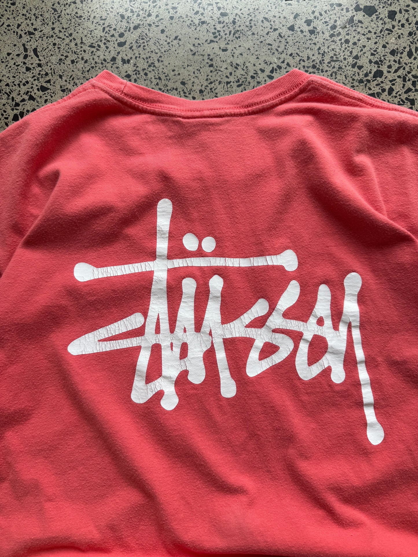 Stussy salmon graphic T-shirt, size L, laid on polished concrete background — bold front logo on soft salmon cotton.