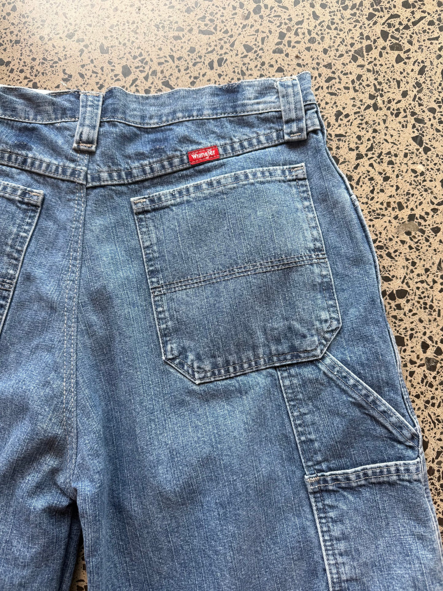 Wrangler mid blue wash denim jorts, size 30W, laid on polished concrete background. Knee cut with classic stitching and soft wash