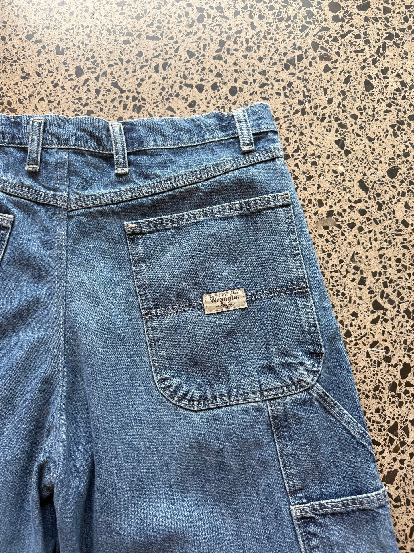 Wrangler Original dark blue wash denim jorts, size 30W, laid on polished concrete background. Knee cut with classic stitching and soft wash