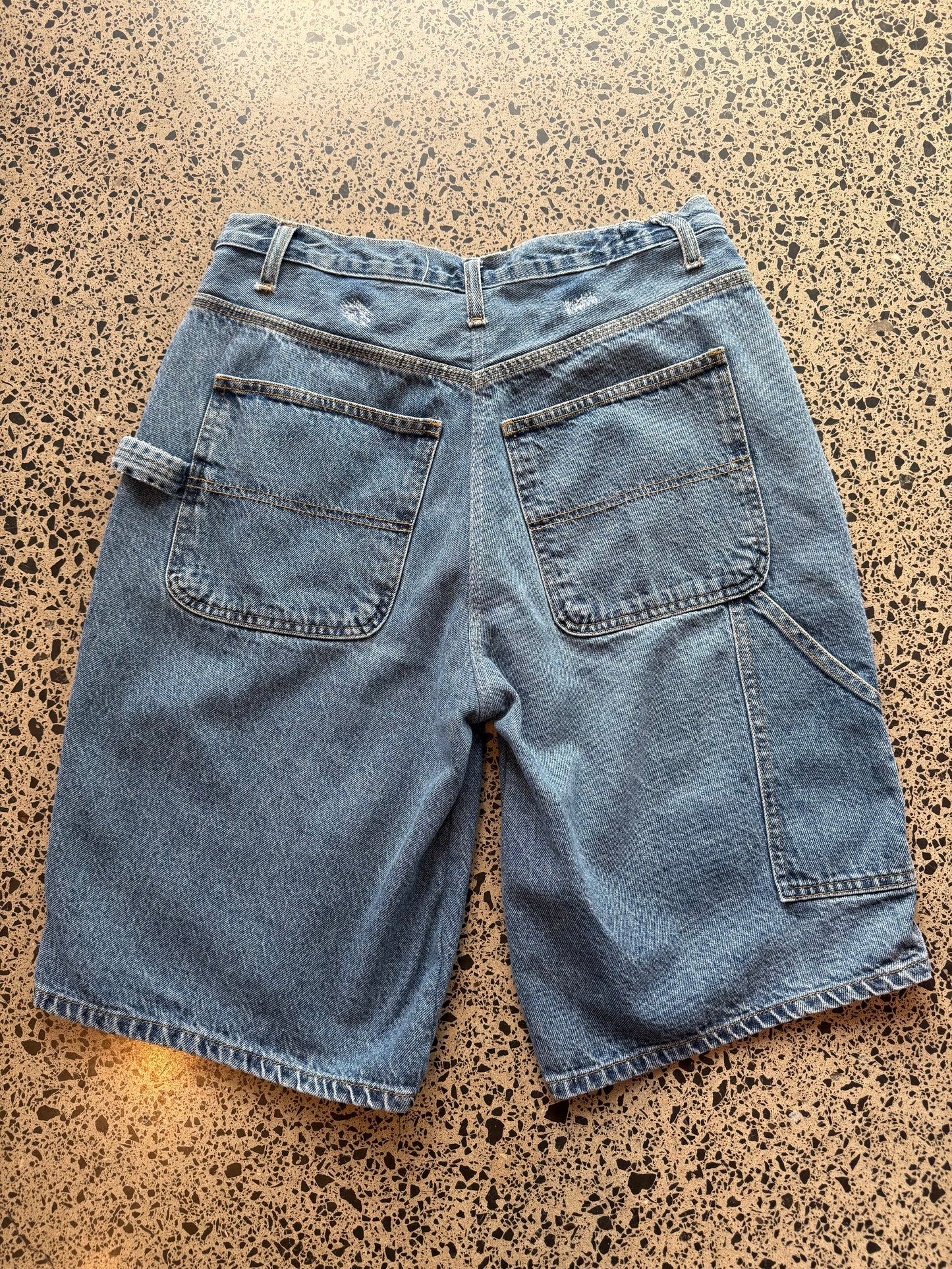 Blue mid-wash denim jorts, size 30W, laid on polished concrete background. Knee cut with classic stitching and soft wash