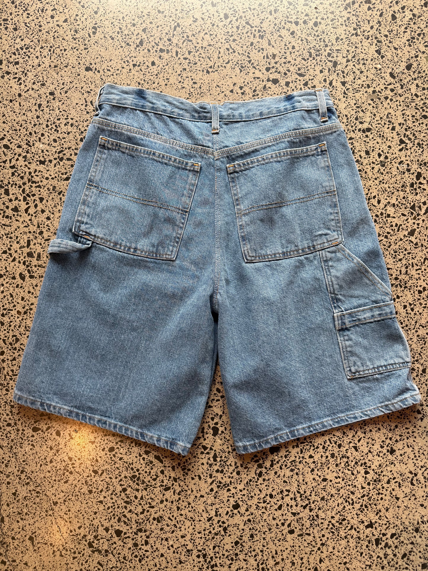 Light - mid blue wash denim jorts, size 32W, laid on polished concrete background. Knee cut with classic stitching and soft wash