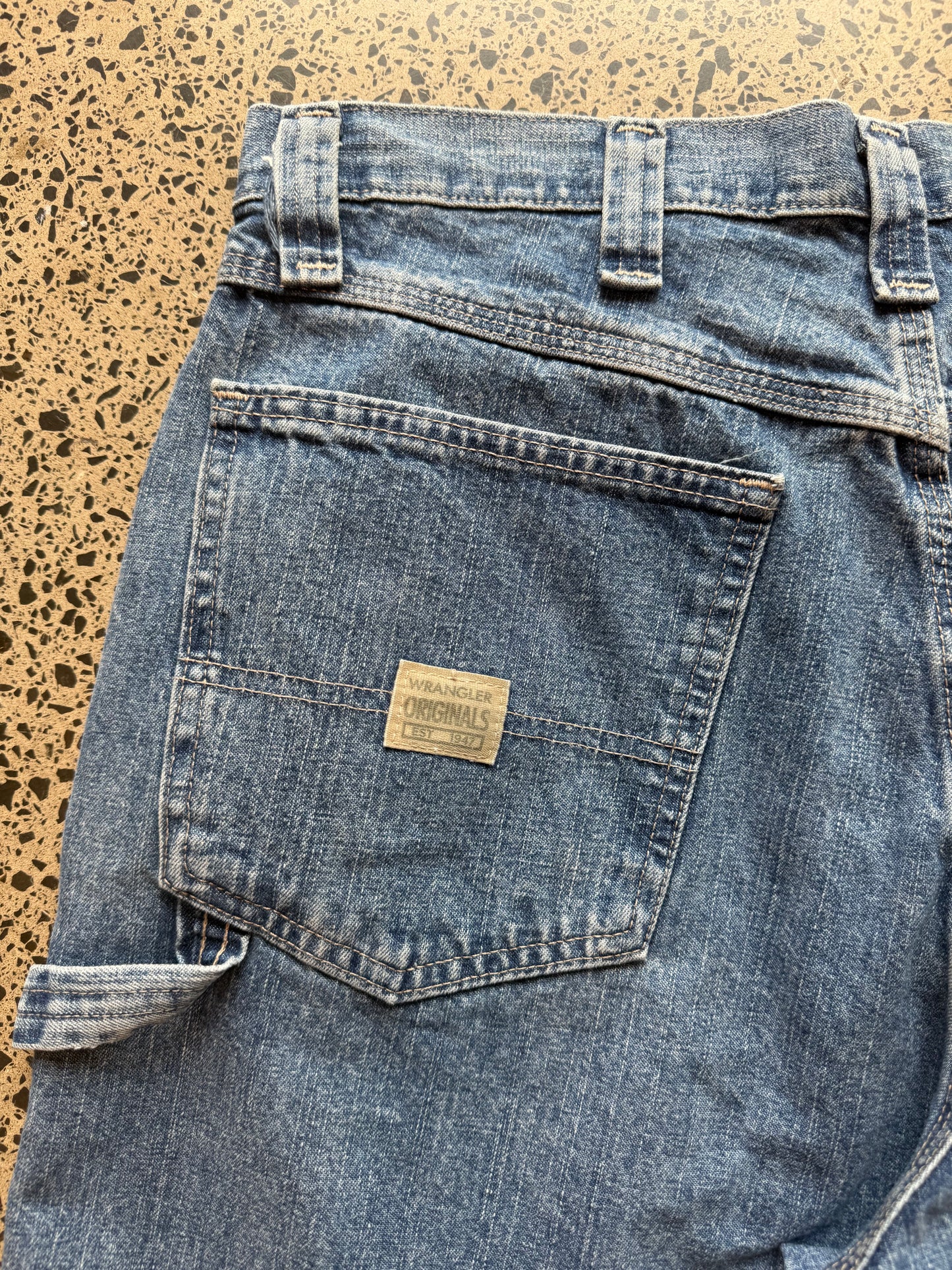 Wrangler Original mid blue wash denim jorts, size 32W, laid on polished concrete background. Knee cut with classic stitching and soft wash