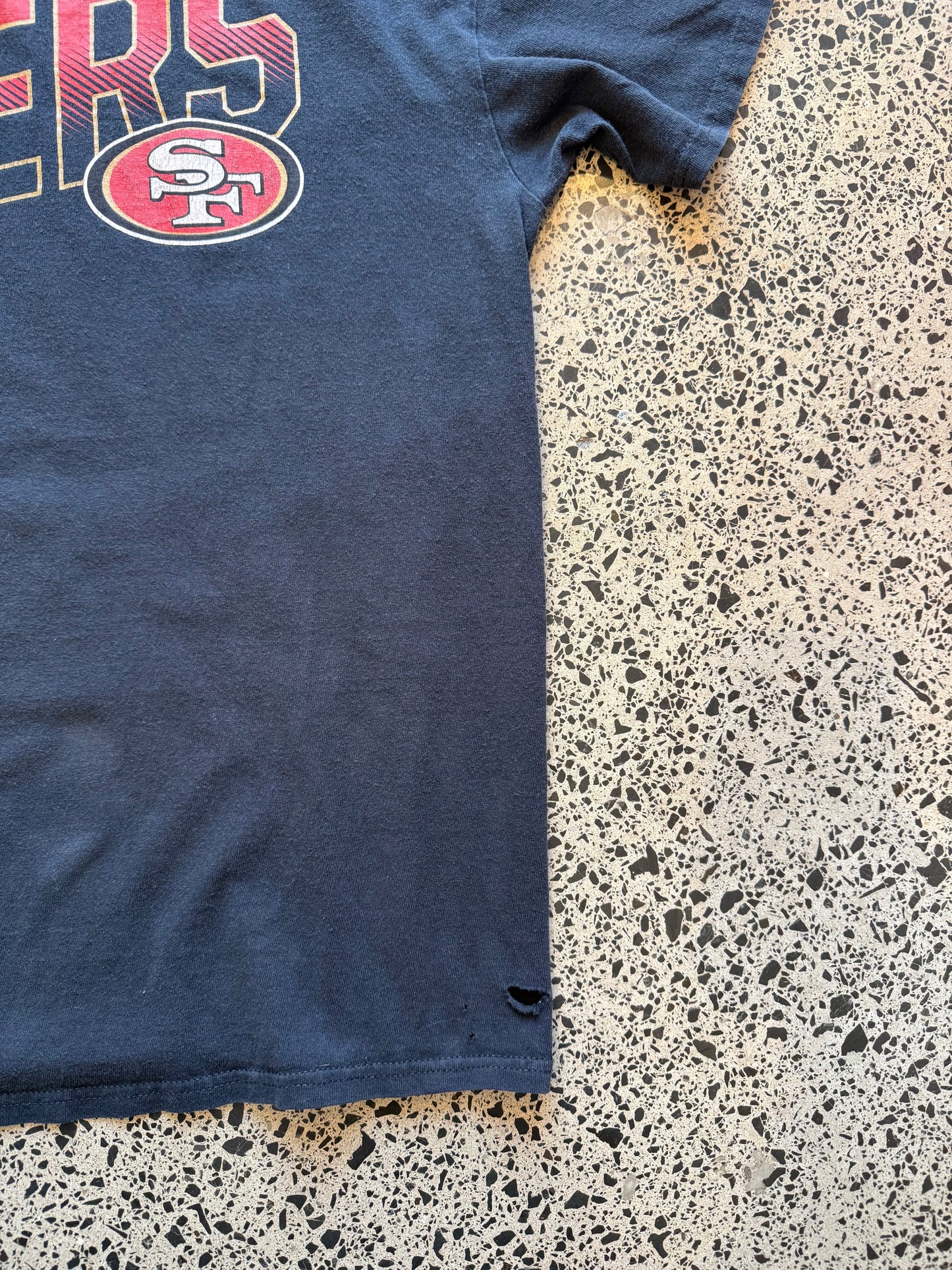 San Francisco 49ers black graphic T-shirt size L laid on polished concrete background — bold red-and-gold team logo on soft cotton.