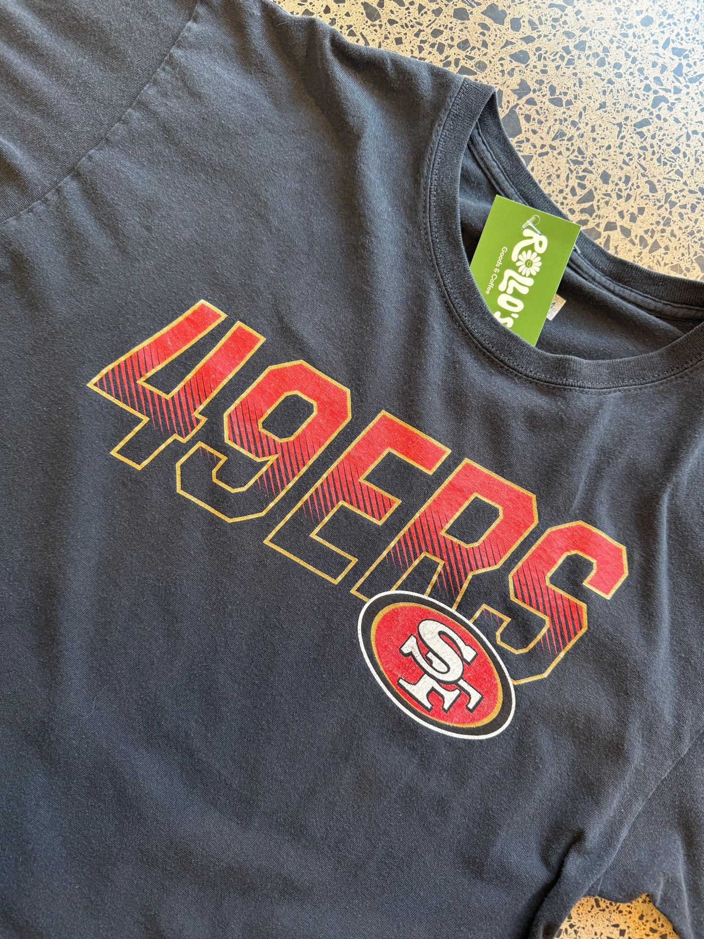 San Francisco 49ers black graphic T-shirt size L laid on polished concrete background — bold red-and-gold team logo on soft cotton.