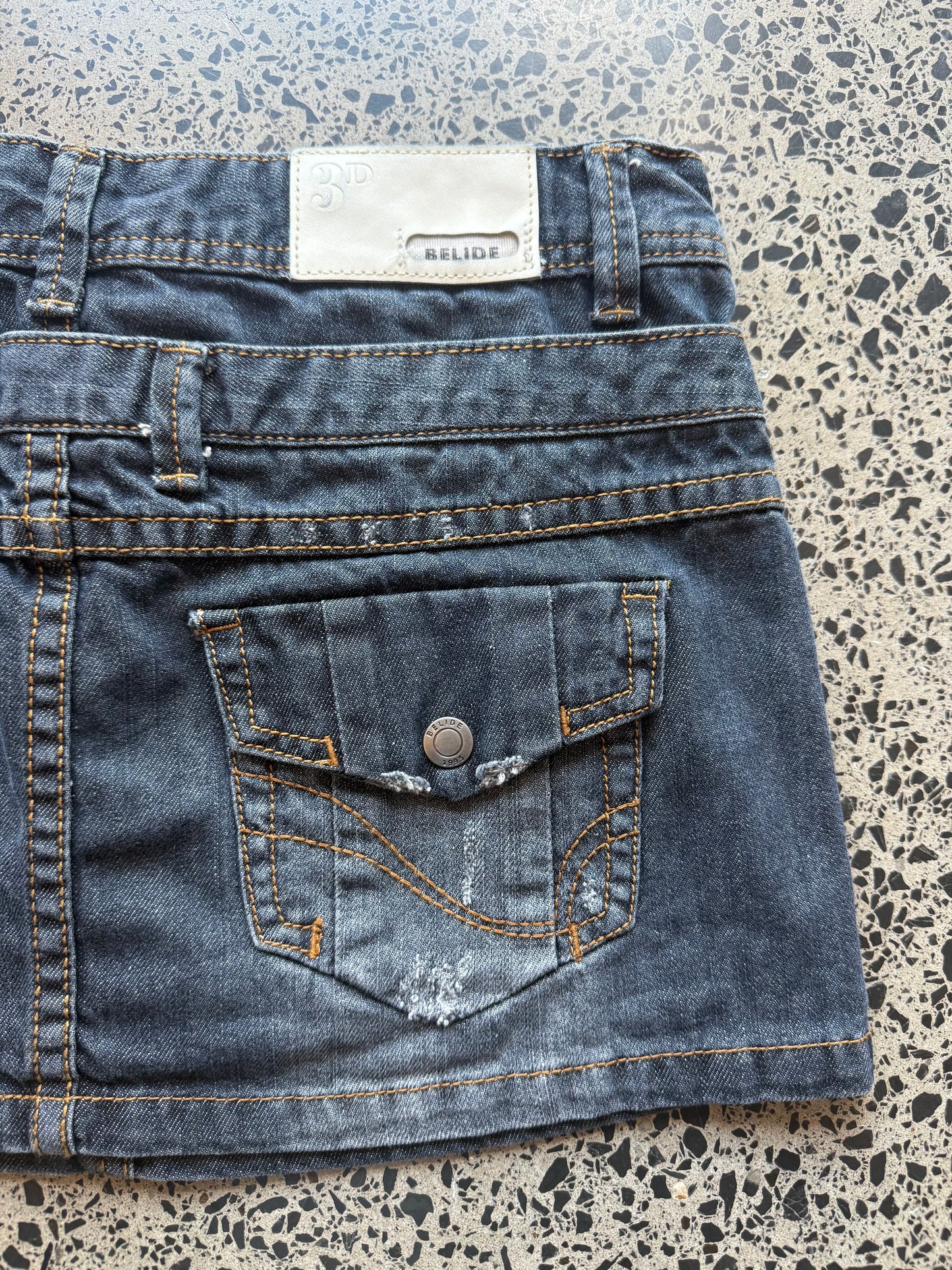 Y2K dark-wash denim mini skirt in XS laid on a polished concrete background — A-line cut with classic stitching and raw hem details.