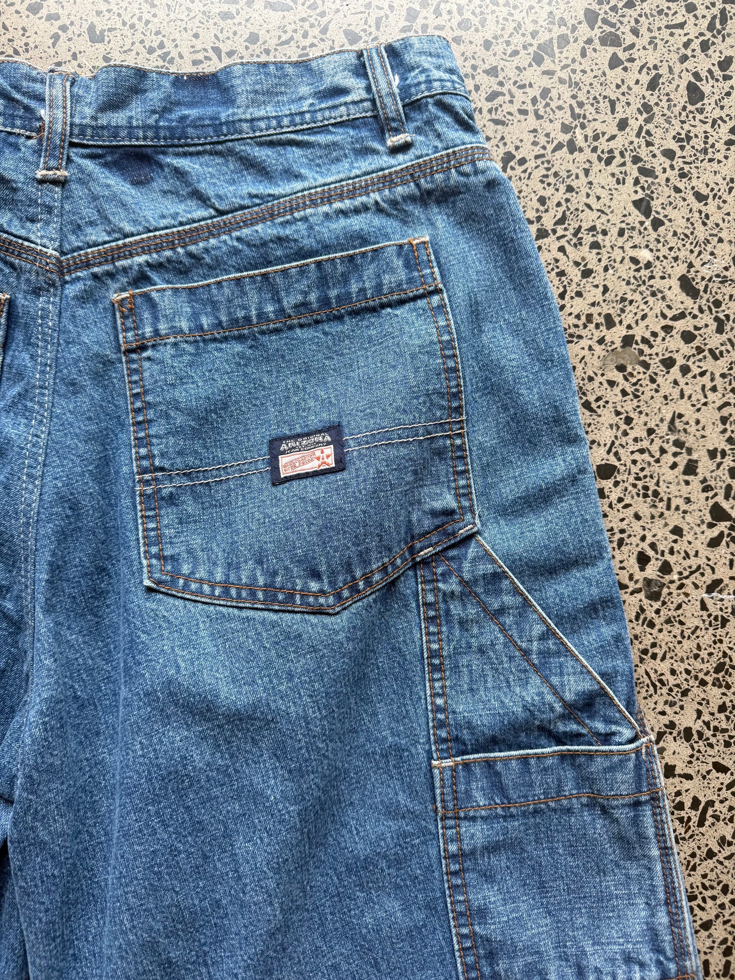 Arizona mid wash denim jorts, size 30W, laid on polished concrete background — below knee cut t with classic stitching and soft faded wash.