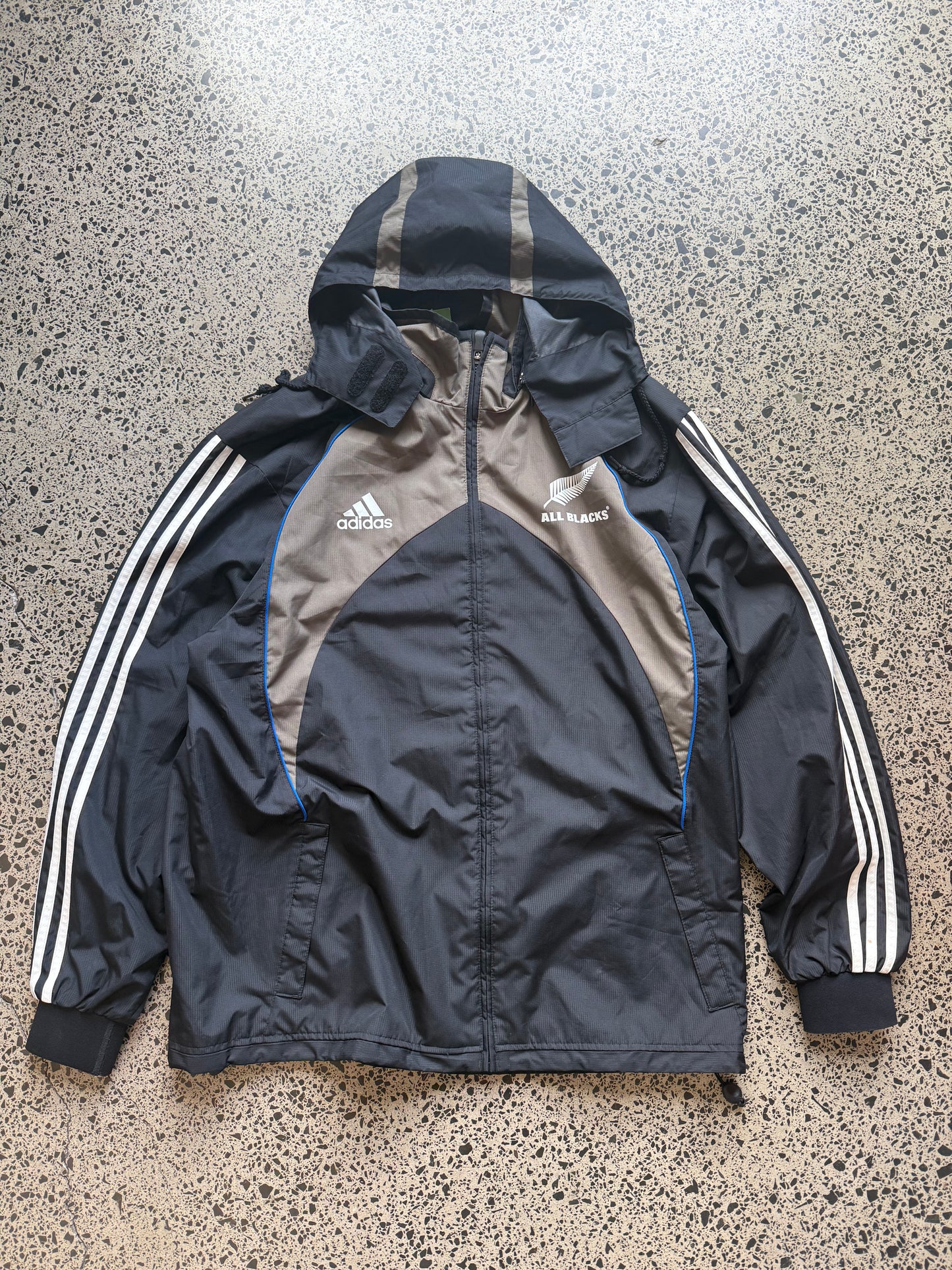 2000s All Blacks rugby jacket size XL laid on polished concrete background — black with silver detailing and All Blacks logo.