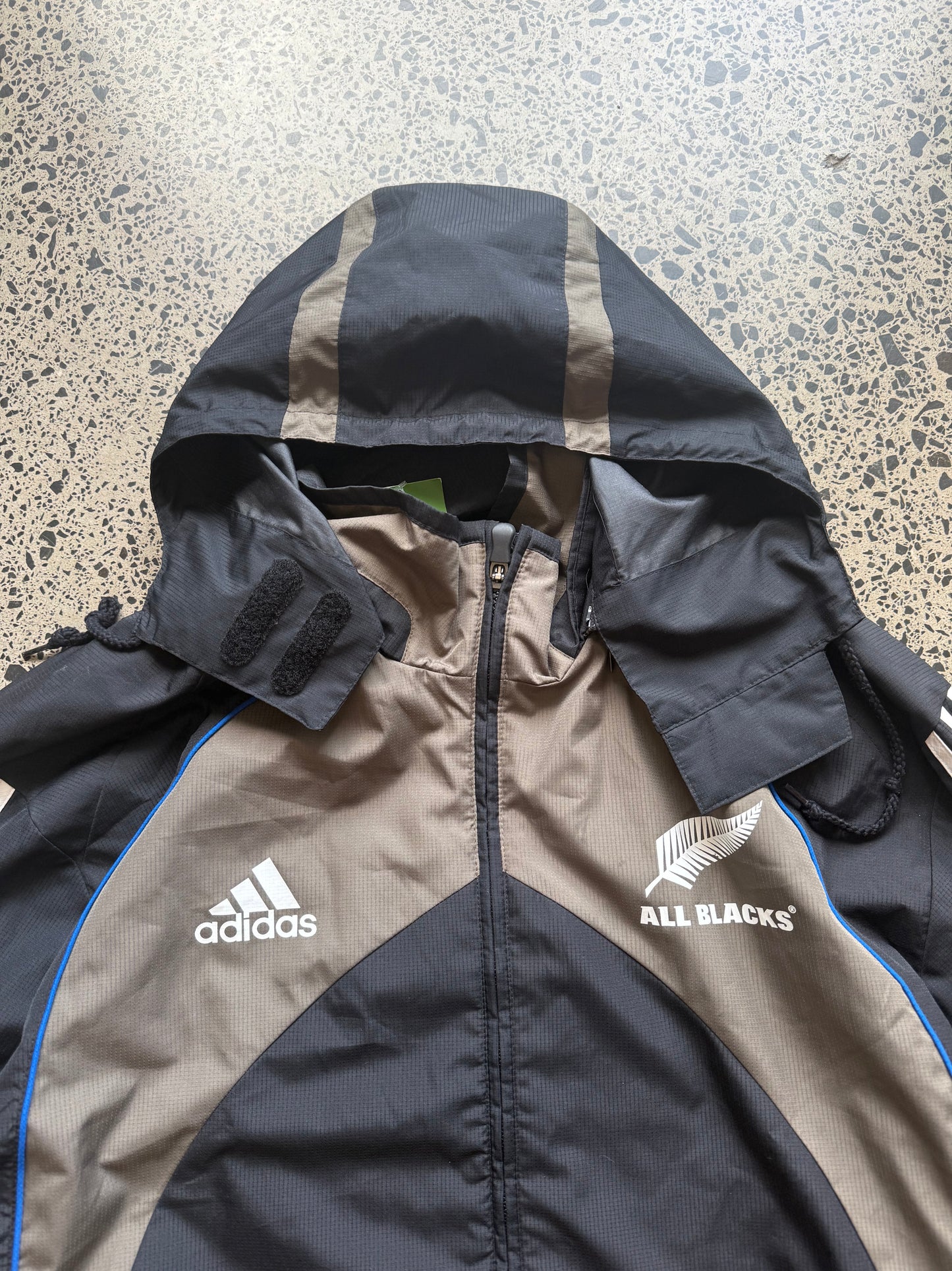 2000s All Blacks rugby jacket size XL laid on polished concrete background — black with silver detailing and All Blacks logo.