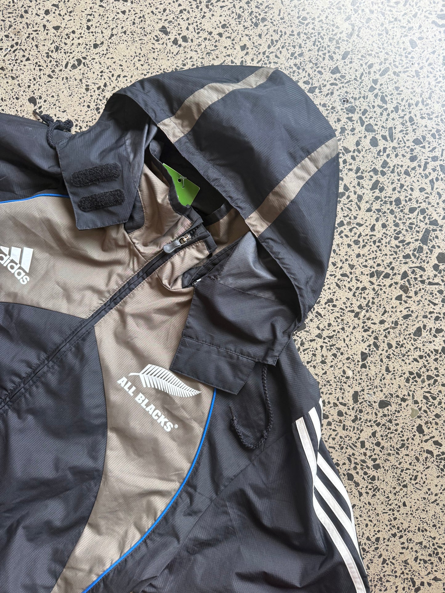 2000s All Blacks rugby jacket size XL laid on polished concrete background — black with silver detailing and All Blacks logo.