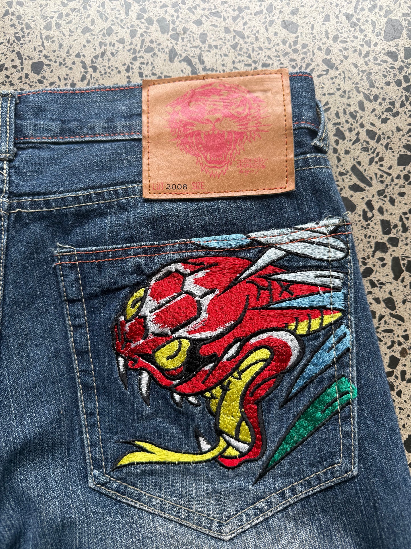 2008 Ed Hardy Snake and Dragon embroidered denim jeans with rhinestone detailing laid flat on a polished concrete background.