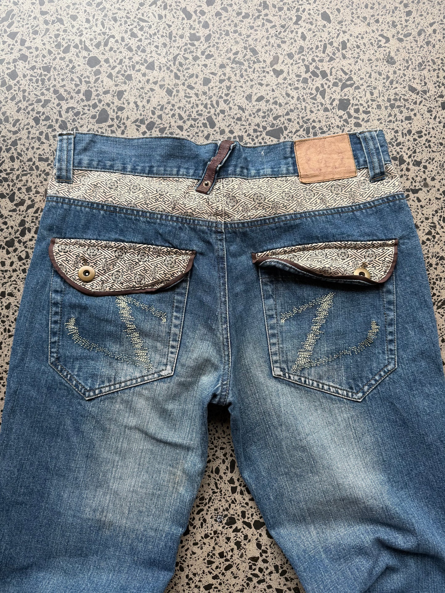 Seku light wash embroidered denim jeans with red and gold dragon detailing laid flat on a polished concrete background.