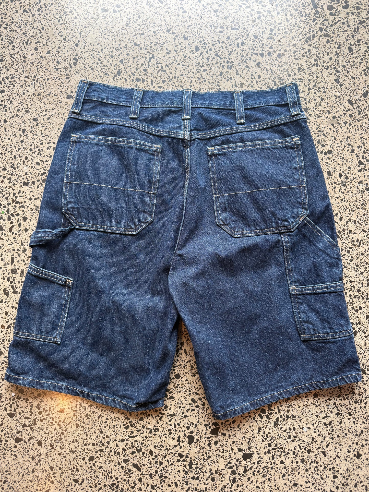 Dark blue wash denim jorts, size 34W, laid on polished concrete background. Knee cut with classic stitching and soft wash