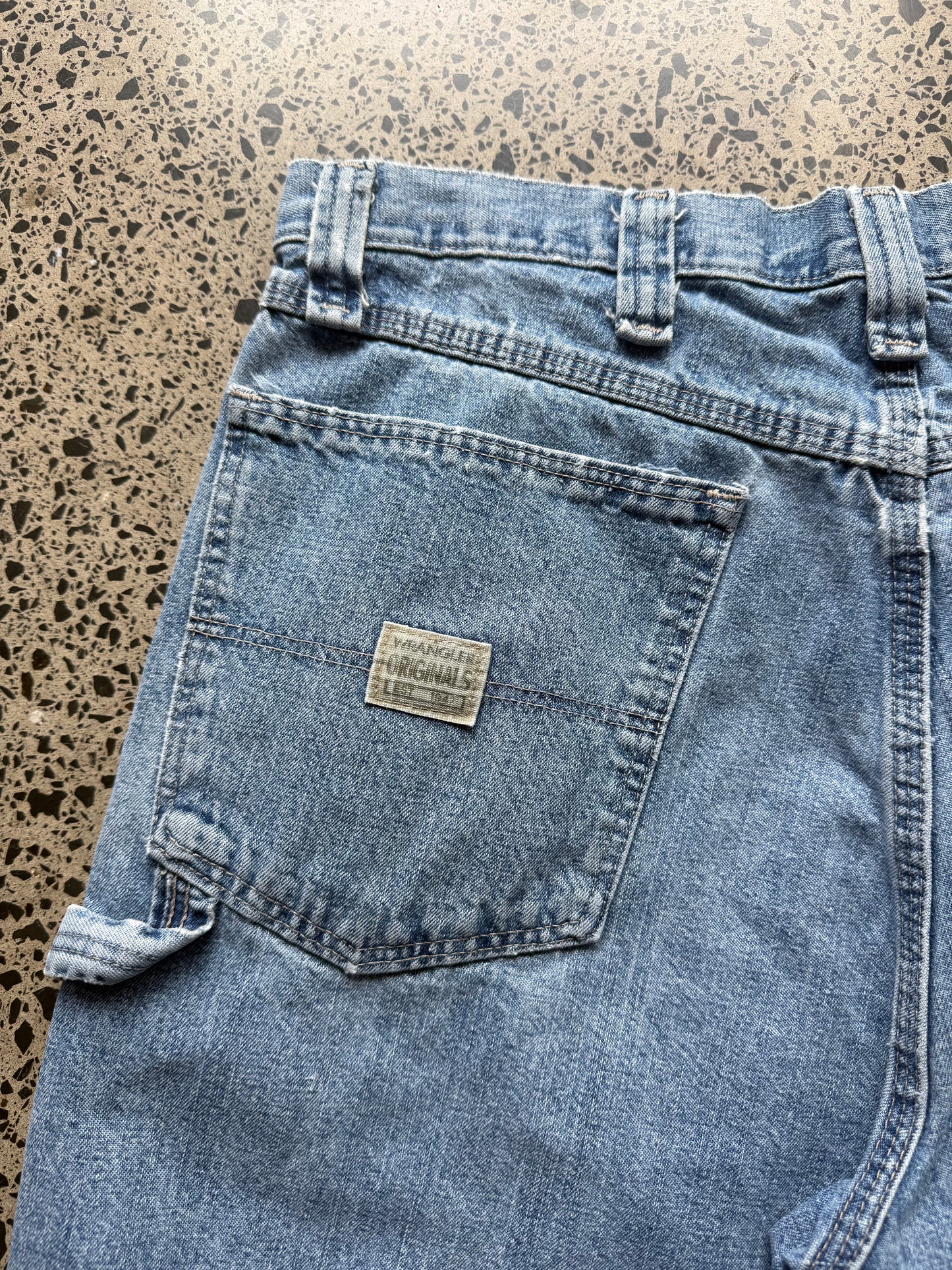 Wrangler original light-wash denim jorts, size 34W, laid on polished concrete background. Knee cut with classic stitching and soft wash
