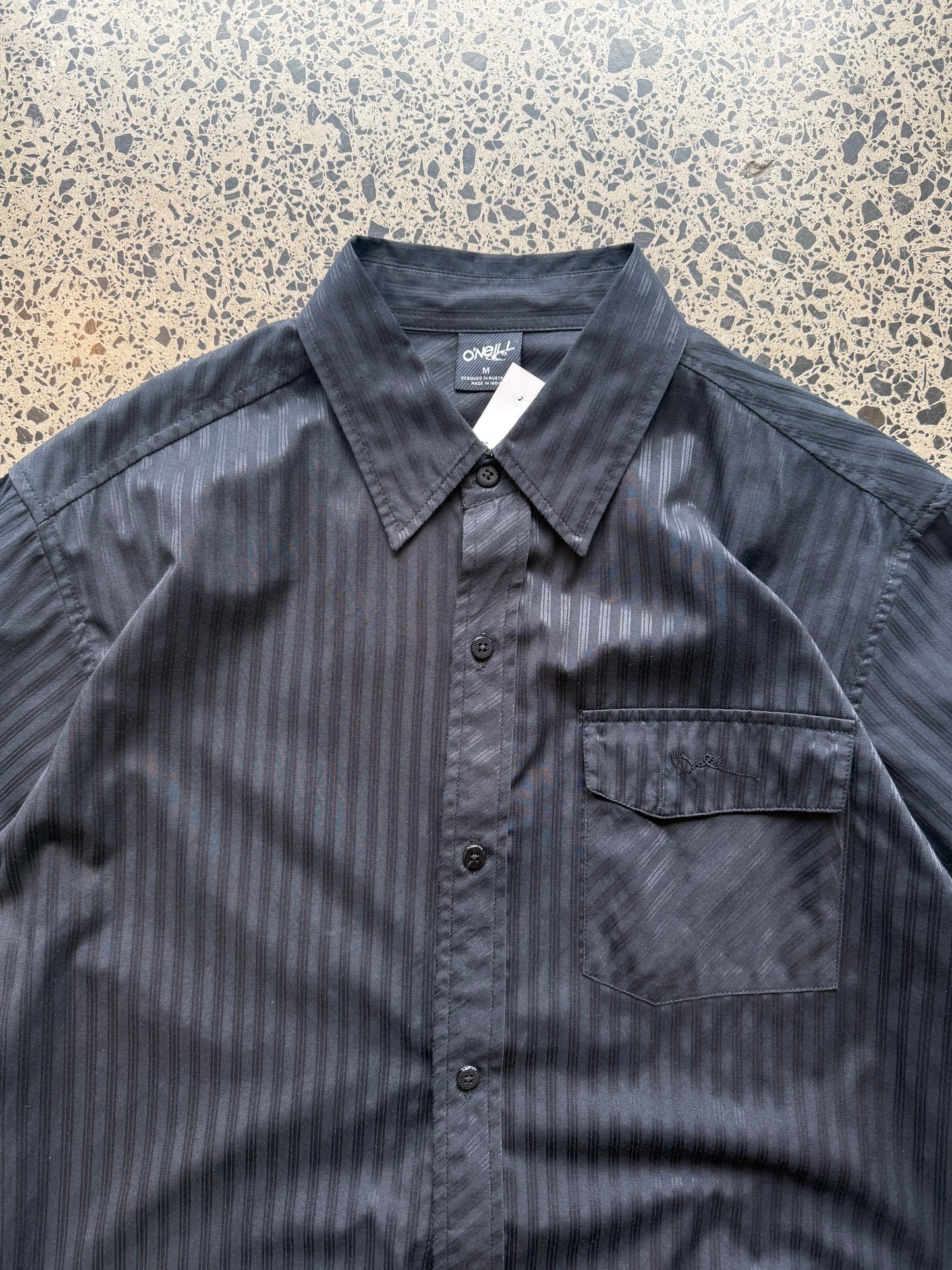 O’Neill short-sleeve button-up shirt in size M laid flat on a polished concrete background featuring relaxed surf-style tailoring