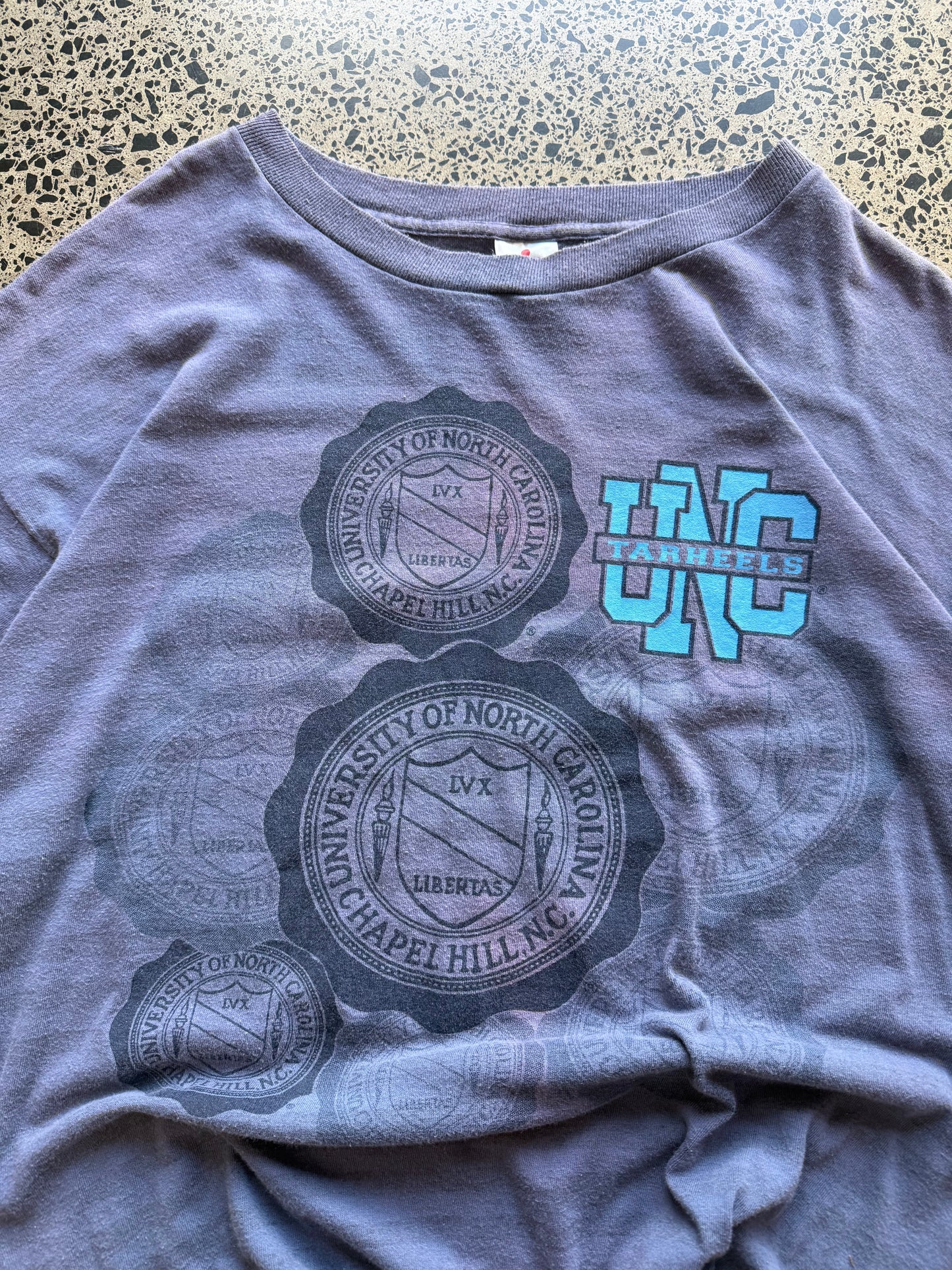 Faded blue 80s North Carolina Tarheels graphic t-shirt with a chest print, size XL, laid flat on a polished concrete background
