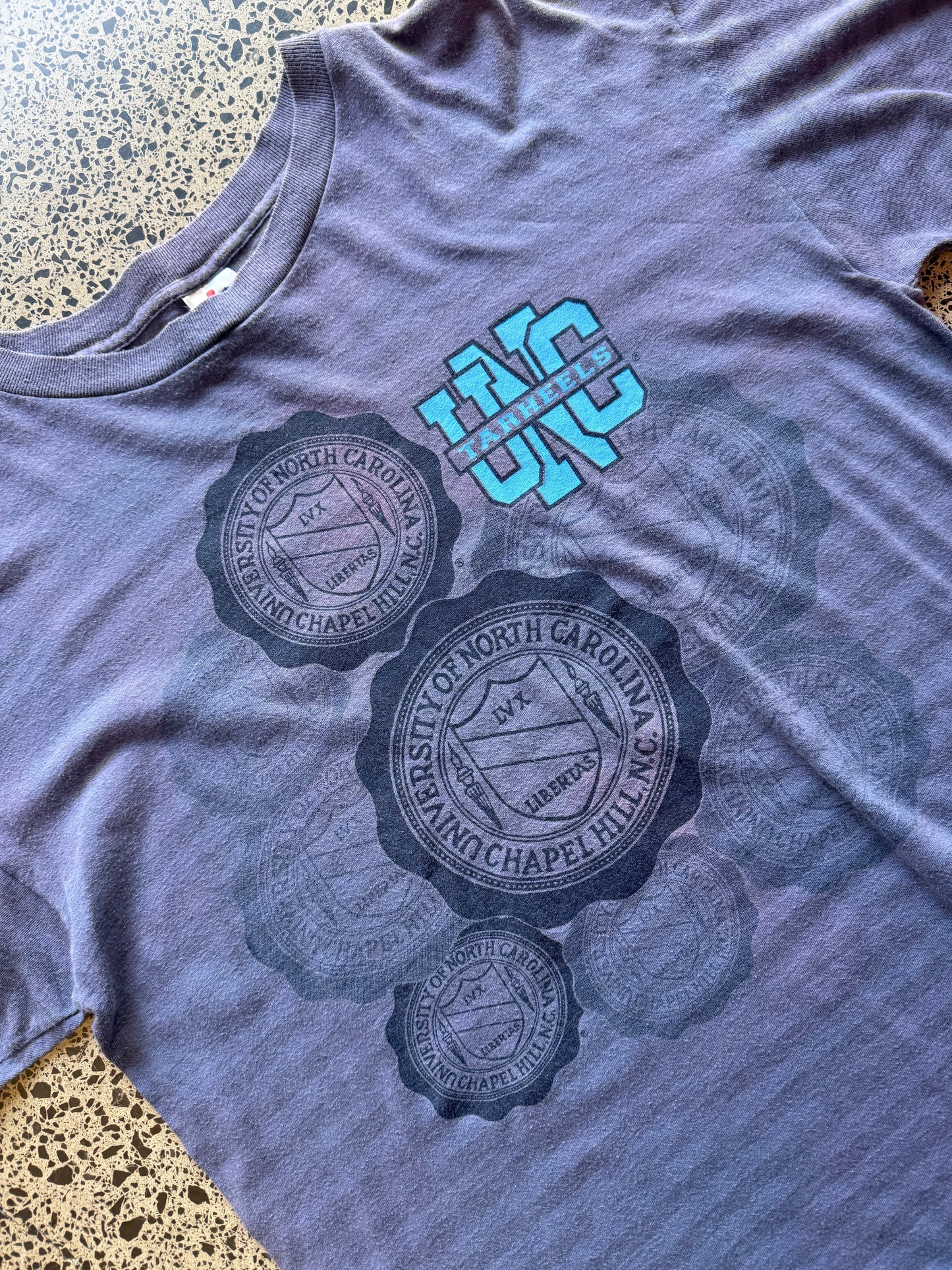 Faded blue 80s North Carolina Tarheels graphic t-shirt with a chest print, size XL, laid flat on a polished concrete background