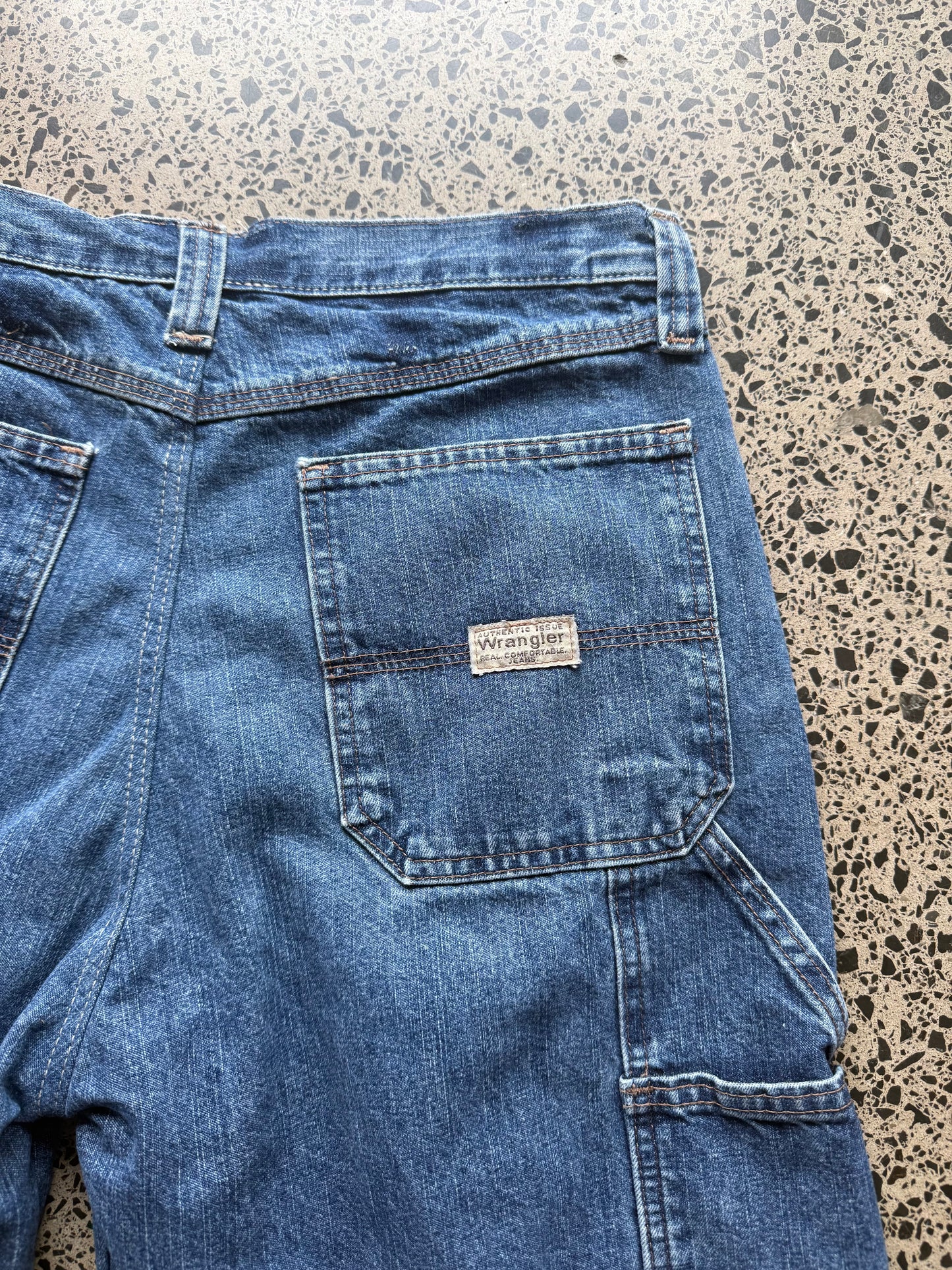 Wrangler Original dark blue wash denim jorts, size 32W, laid on polished concrete background. Knee cut with classic stitching and soft wash