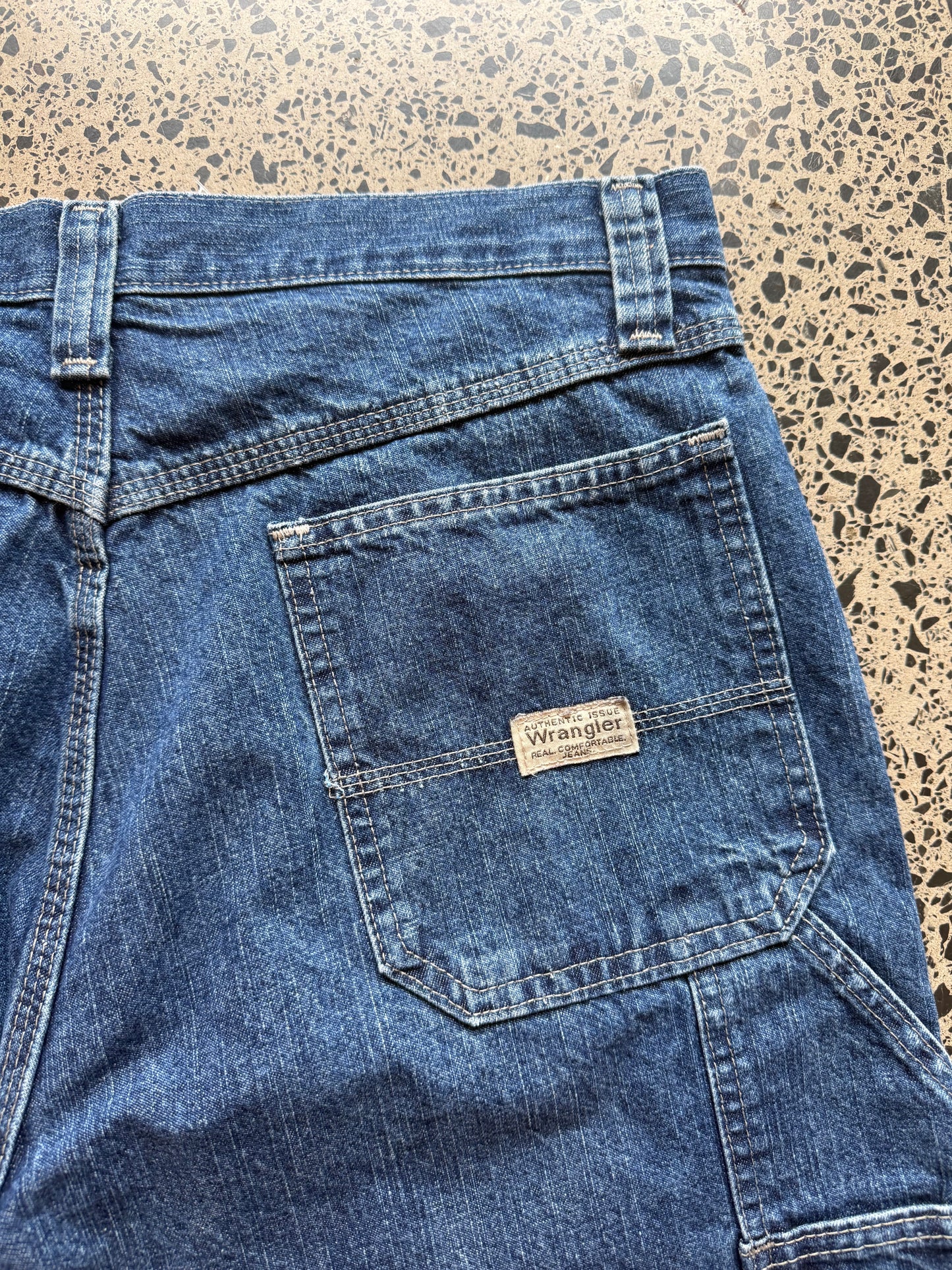 Wrangler Original dark blue wash denim jorts, size 32W, laid on polished concrete background. Knee cut with classic stitching and soft wash