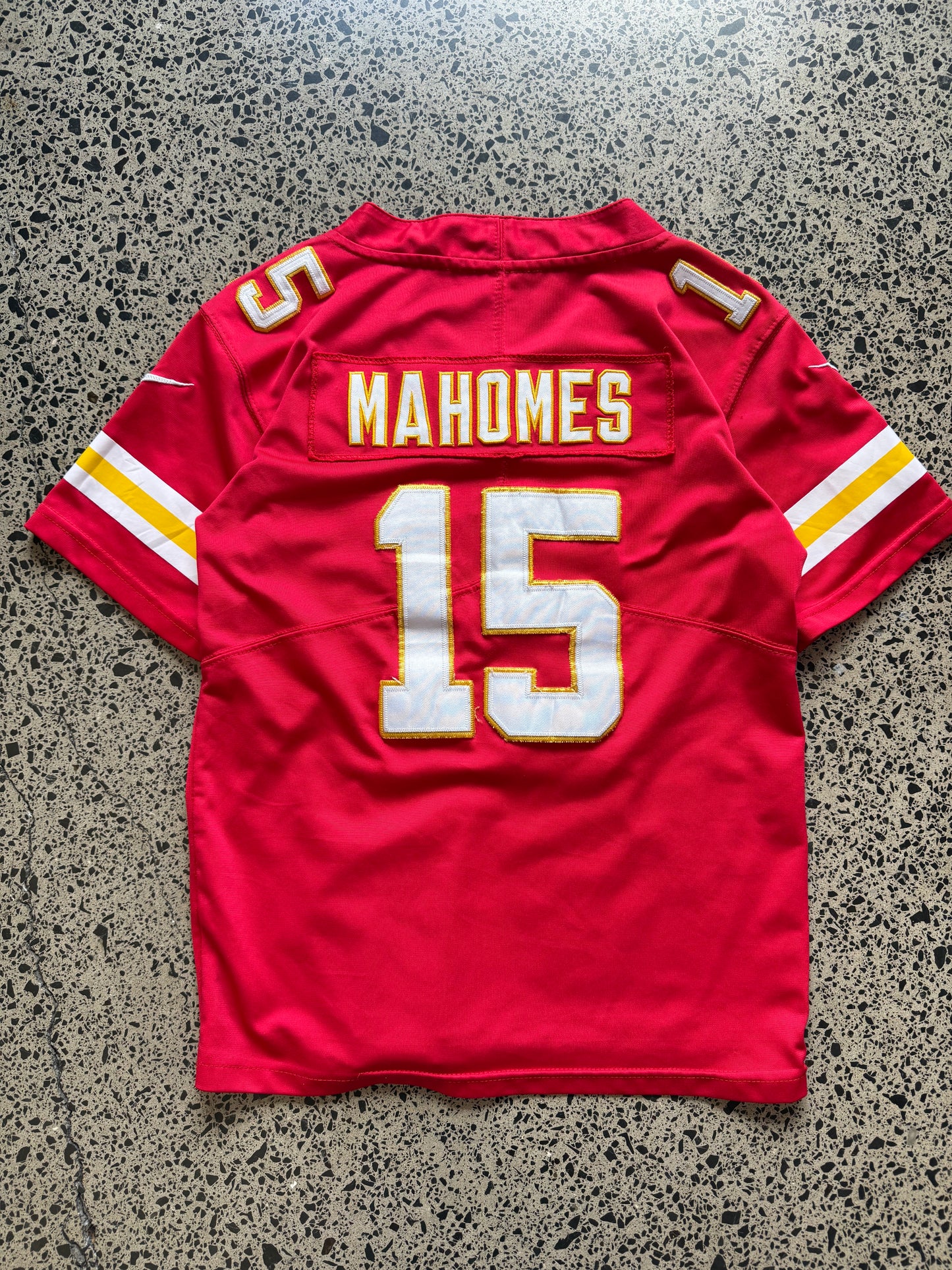 Kansas City Chiefs Patrick Mahomes NFL jersey in Youth Large laid on polished concrete background — red with white and yellow detailing and Chiefs logo.