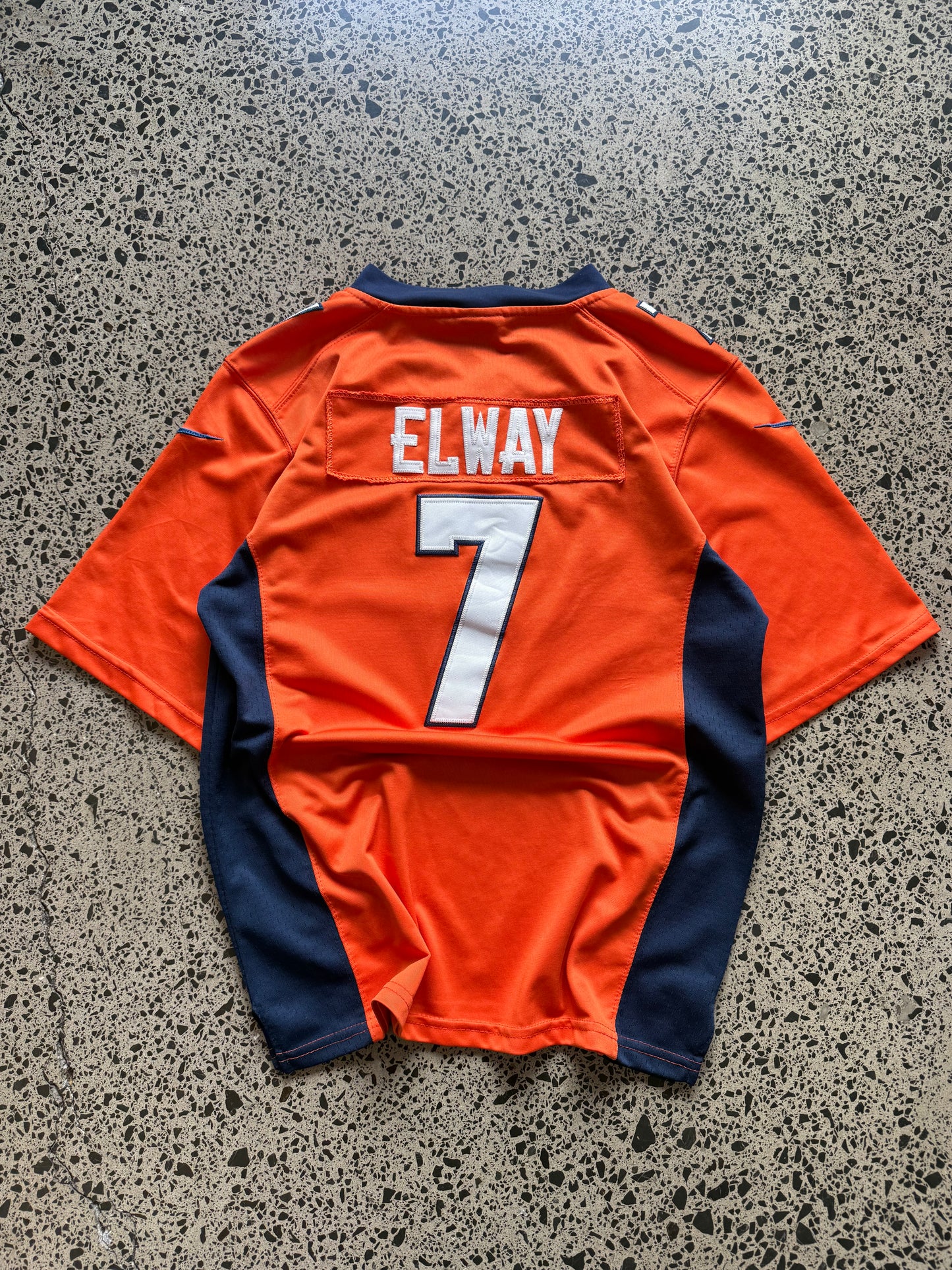 Denver Broncos John Elway NFL jersey size L laid on polished concrete background — bright orange with navy and white detailing and Broncos logo.