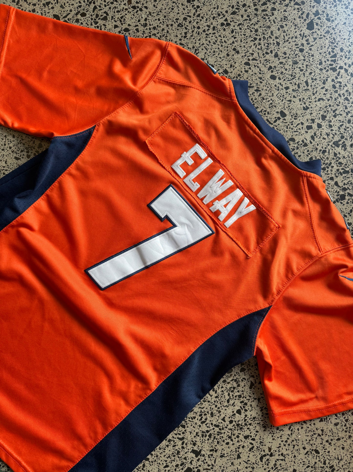 Denver Broncos John Elway NFL jersey size L laid on polished concrete background — bright orange with navy and white detailing and Broncos logo.