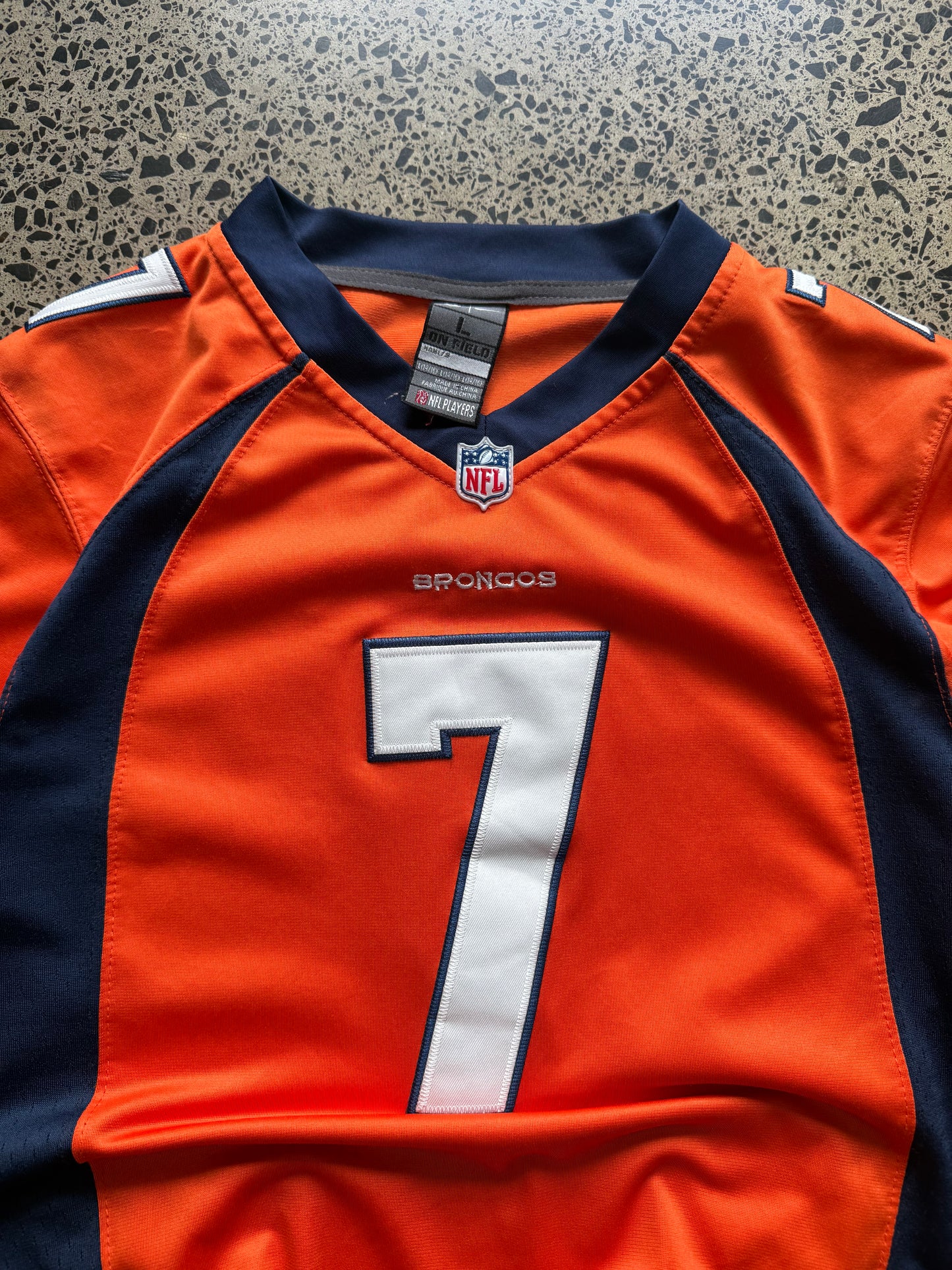 Denver Broncos John Elway NFL jersey size L laid on polished concrete background — bright orange with navy and white detailing and Broncos logo.