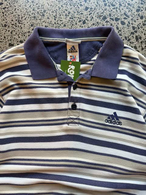 Vintage striped Adidas collared shirt laid flat on polished concrete background with classic stripe pattern and embroidered Adidas Trefoil logo.
