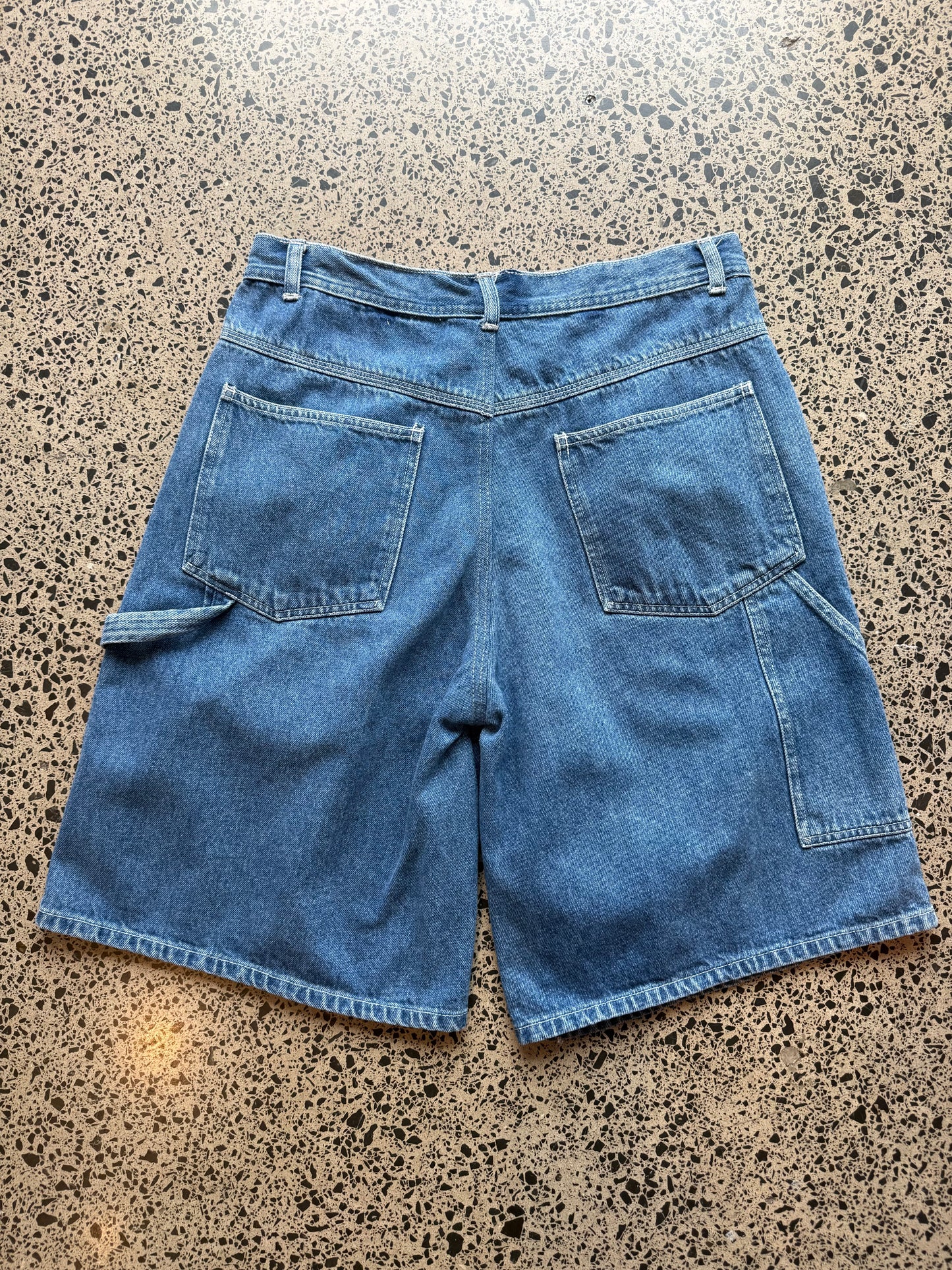 Mid-wash blue wash denim jorts, size 32W, laid on polished concrete background. Knee cut with classic stitching and soft wash