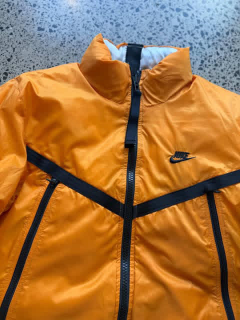 Reversible orange and white Nike jacket laid flat on a polished concrete background featuring classic Nike branding and dual-colour design
