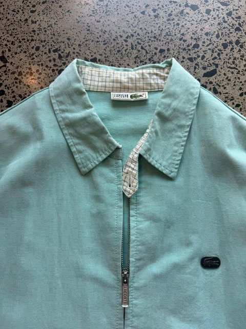 Turquoise Lacoste collared zip-up jacket laid flat on polished concrete background, vintage designer jacket with embroidered crocodile logo and clean fit.