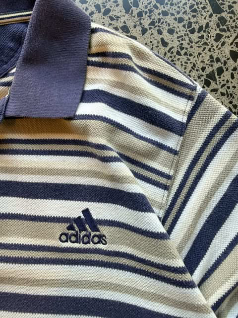 Vintage striped Adidas collared shirt laid flat on polished concrete background with classic stripe pattern and embroidered Adidas Trefoil logo.
