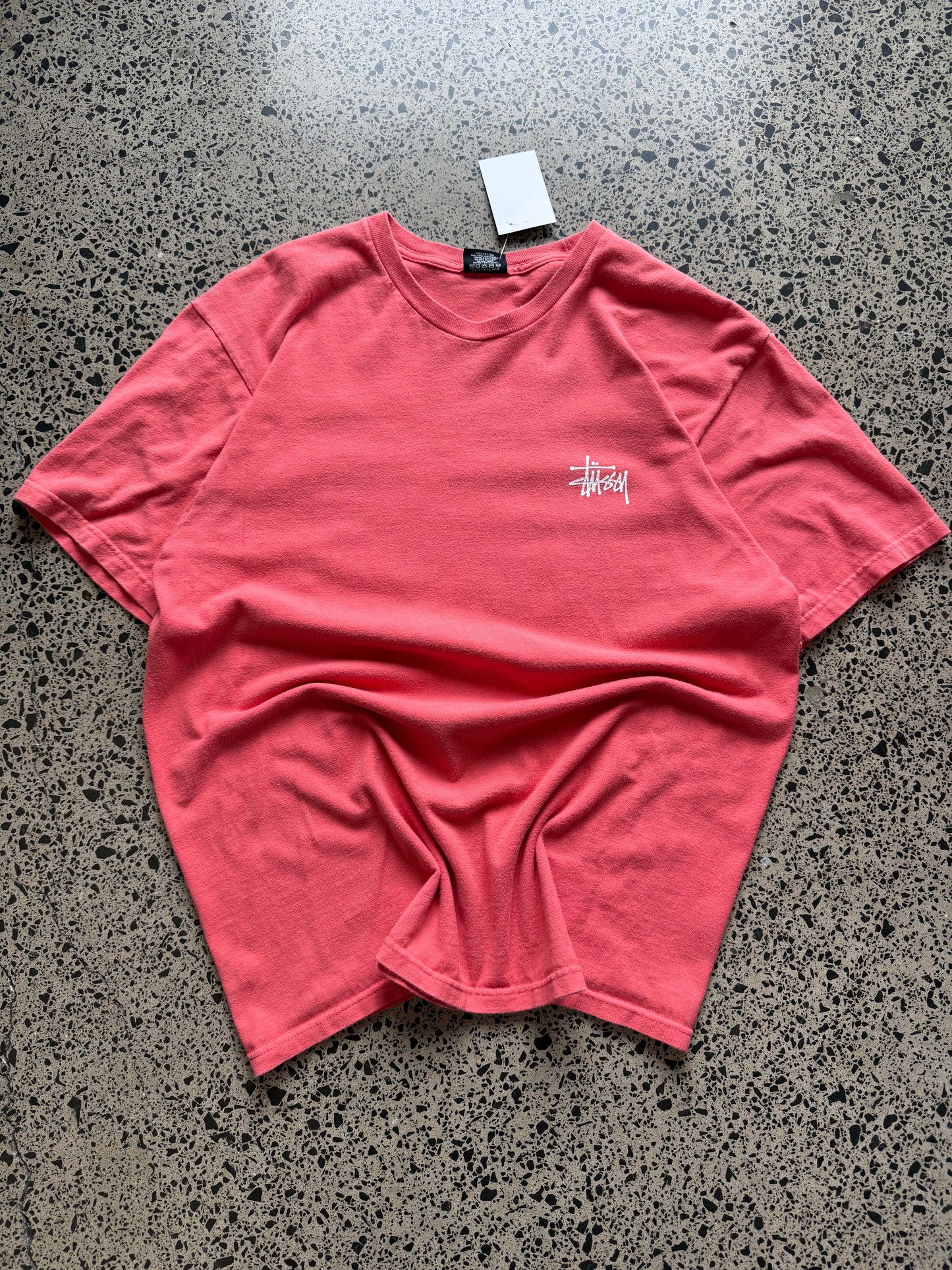 Stussy salmon graphic T-shirt, size L, laid on polished concrete background — bold front logo on soft salmon cotton.
