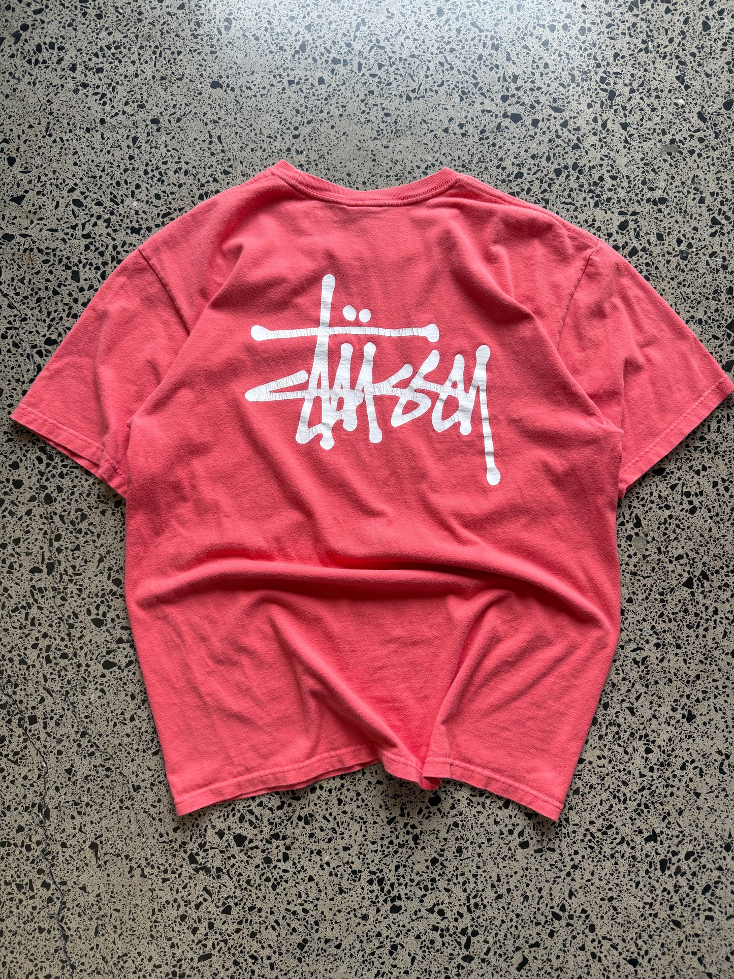 Stussy salmon graphic T-shirt, size L, laid on polished concrete background — bold front logo on soft salmon cotton.