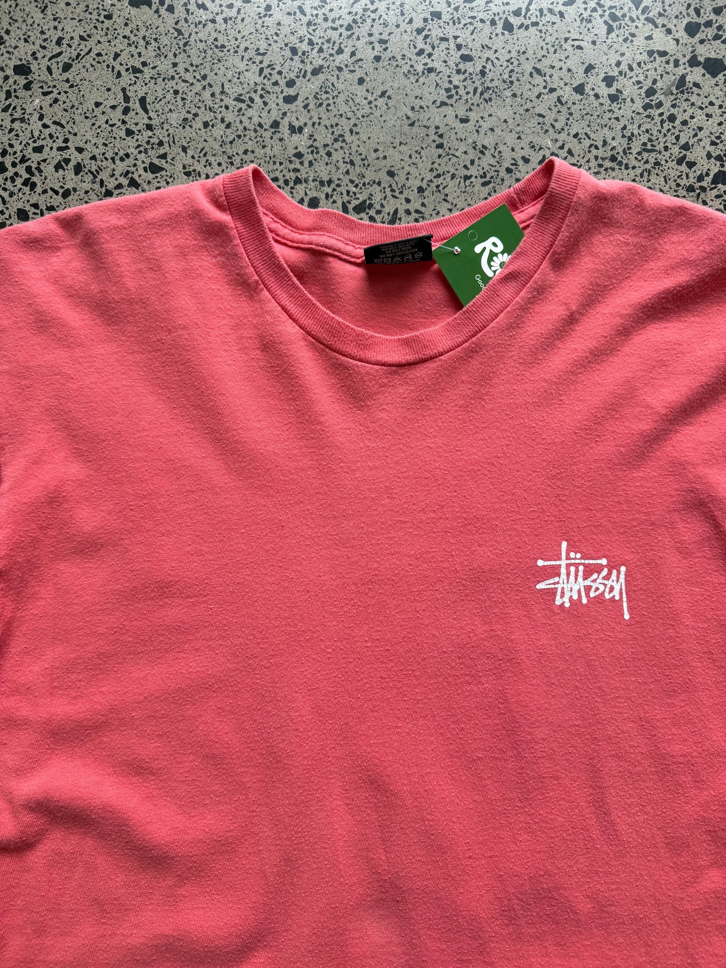 Stussy salmon graphic T-shirt, size L, laid on polished concrete background — bold front logo on soft salmon cotton.