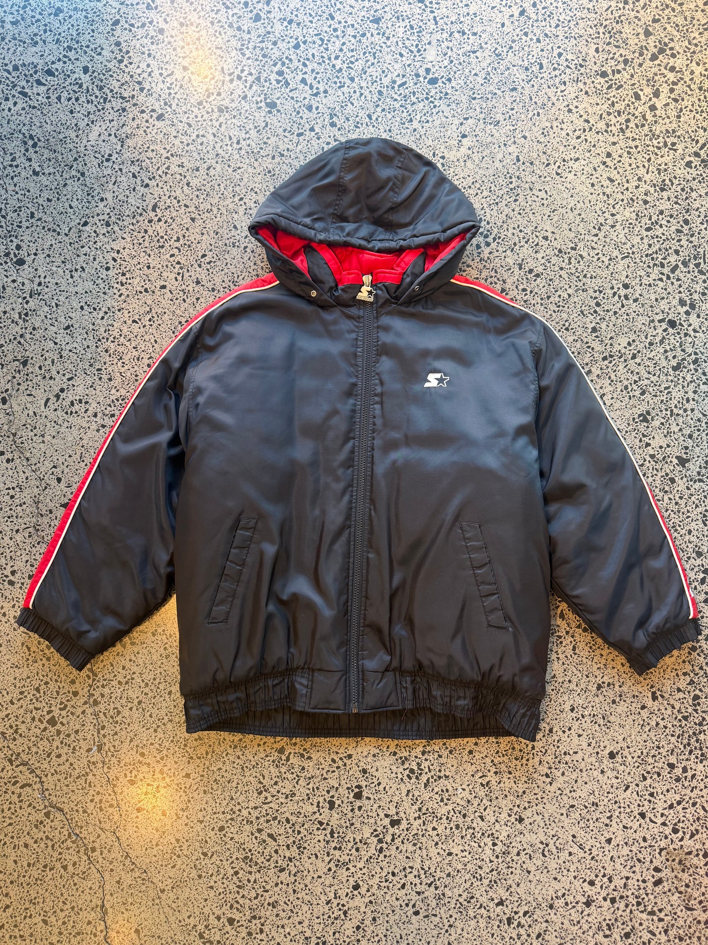 90s black and red Starter jacket size M laid flat on polished concrete background, vintage jacket with bold colour blocking and classic Starter fit.