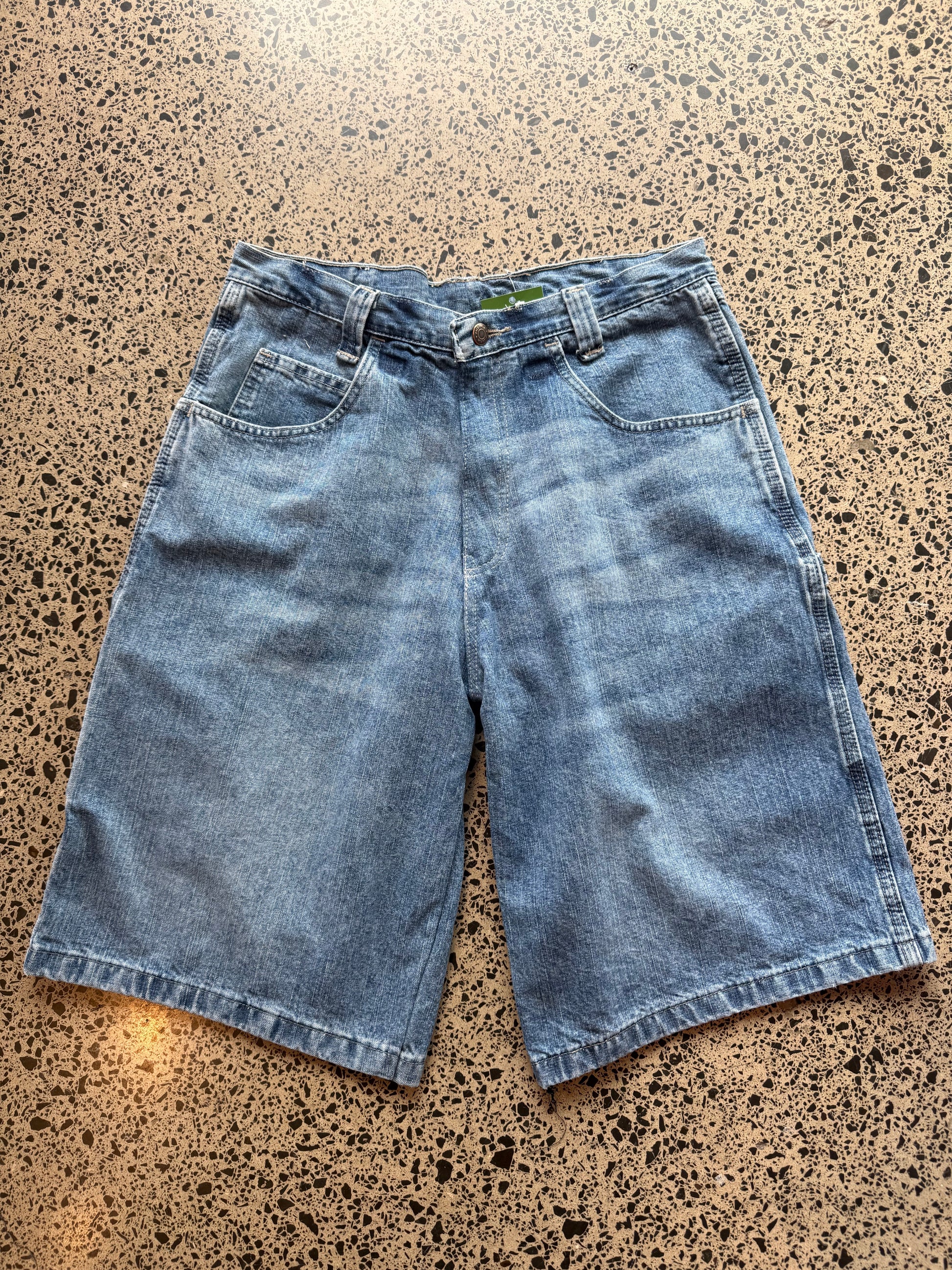 Wrangler mid blue wash denim jorts, size 30W, laid on polished concrete background. Knee cut with classic stitching and soft wash