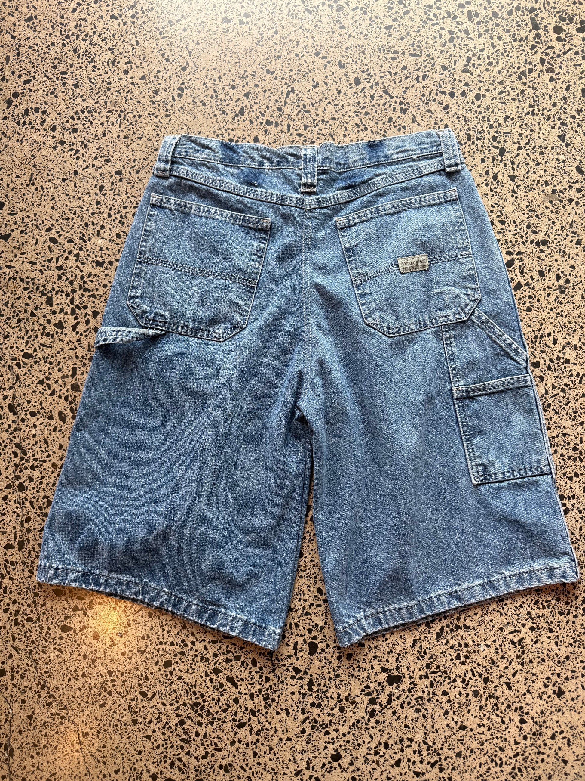 Wrangler mid blue wash denim jorts, size 30W, laid on polished concrete background. Knee cut with classic stitching and soft wash