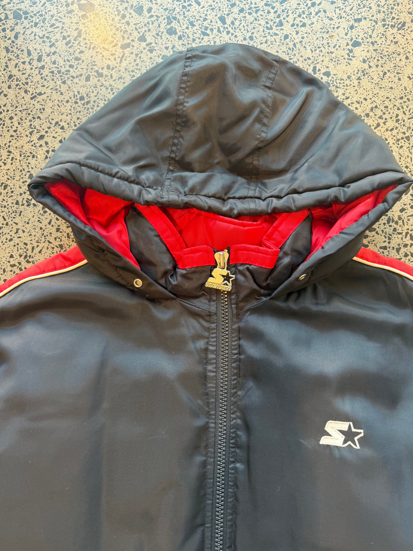 90s black and red Starter jacket size M laid flat on polished concrete background, vintage jacket with bold colour blocking and classic Starter fit.