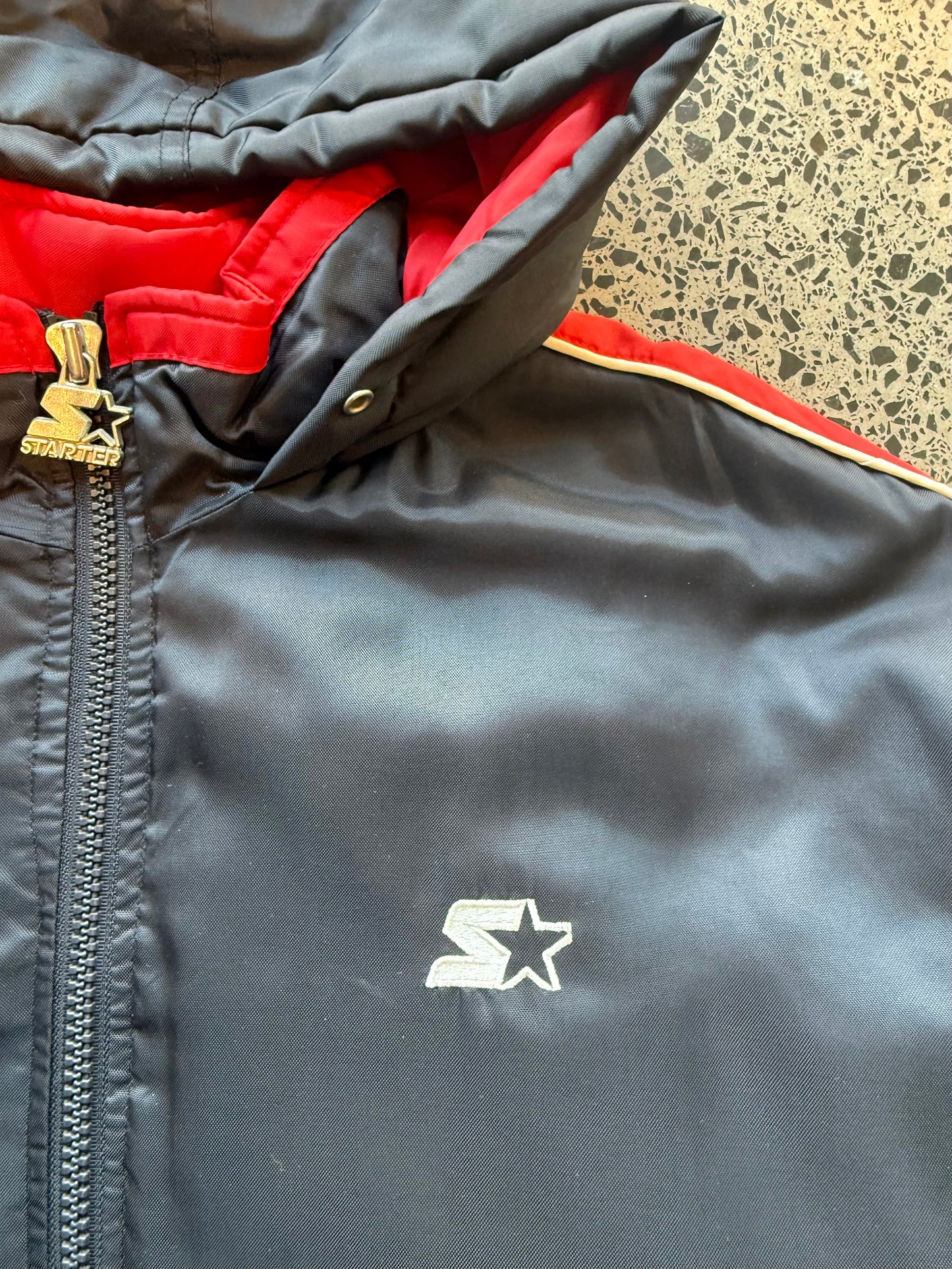 90s black and red Starter jacket size M laid flat on polished concrete background, vintage jacket with bold colour blocking and classic Starter fit.