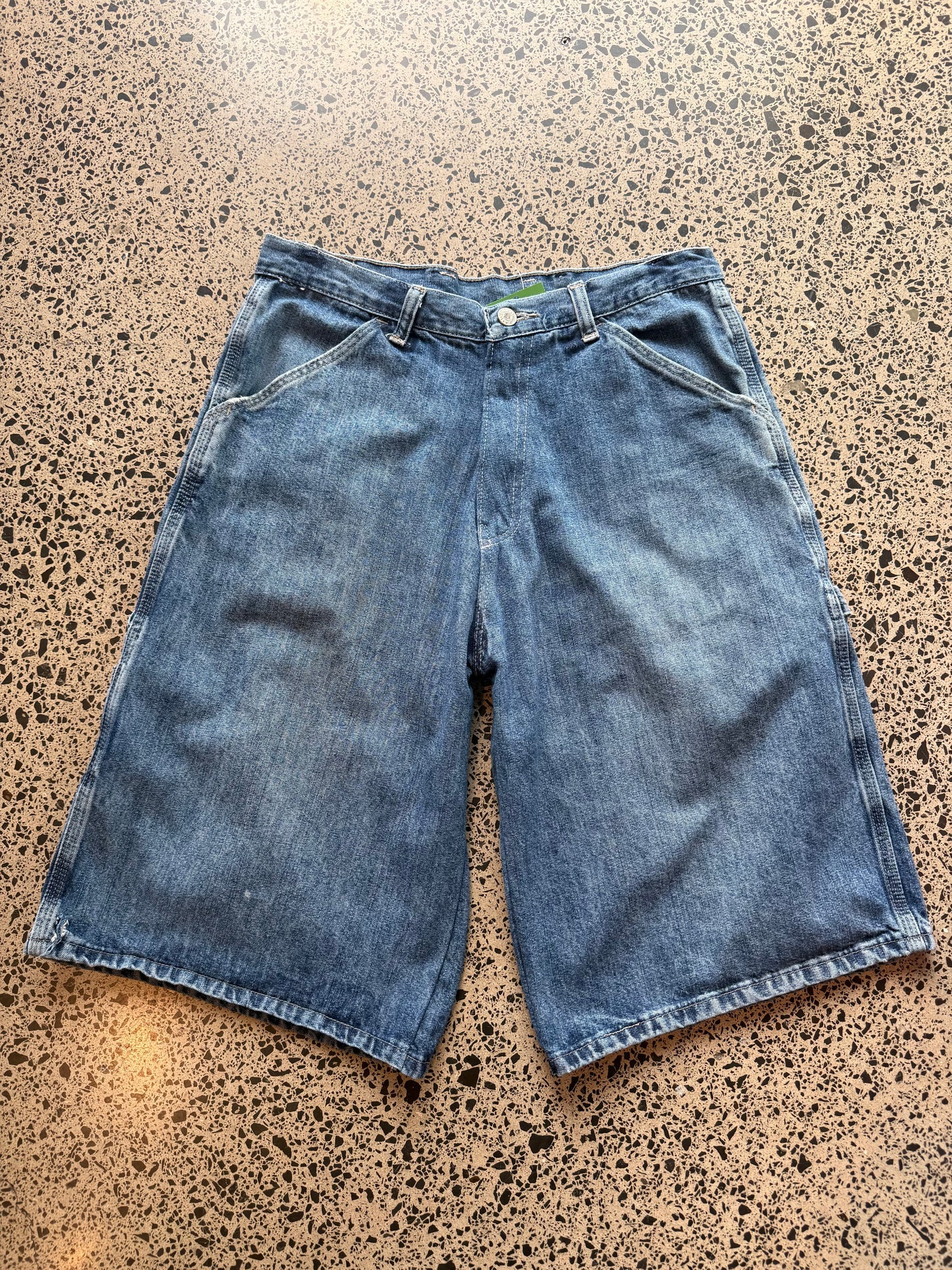 Wrangler Original dark blue wash denim jorts, size 30W, laid on polished concrete background. Knee cut with classic stitching and soft wash