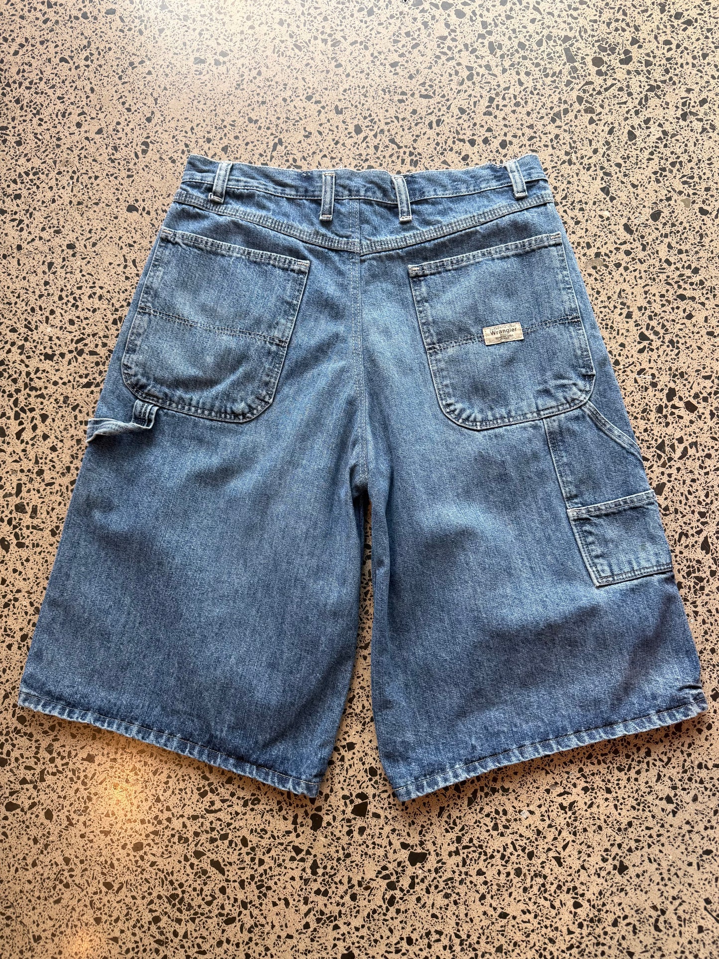 Wrangler Original dark blue wash denim jorts, size 30W, laid on polished concrete background. Knee cut with classic stitching and soft wash