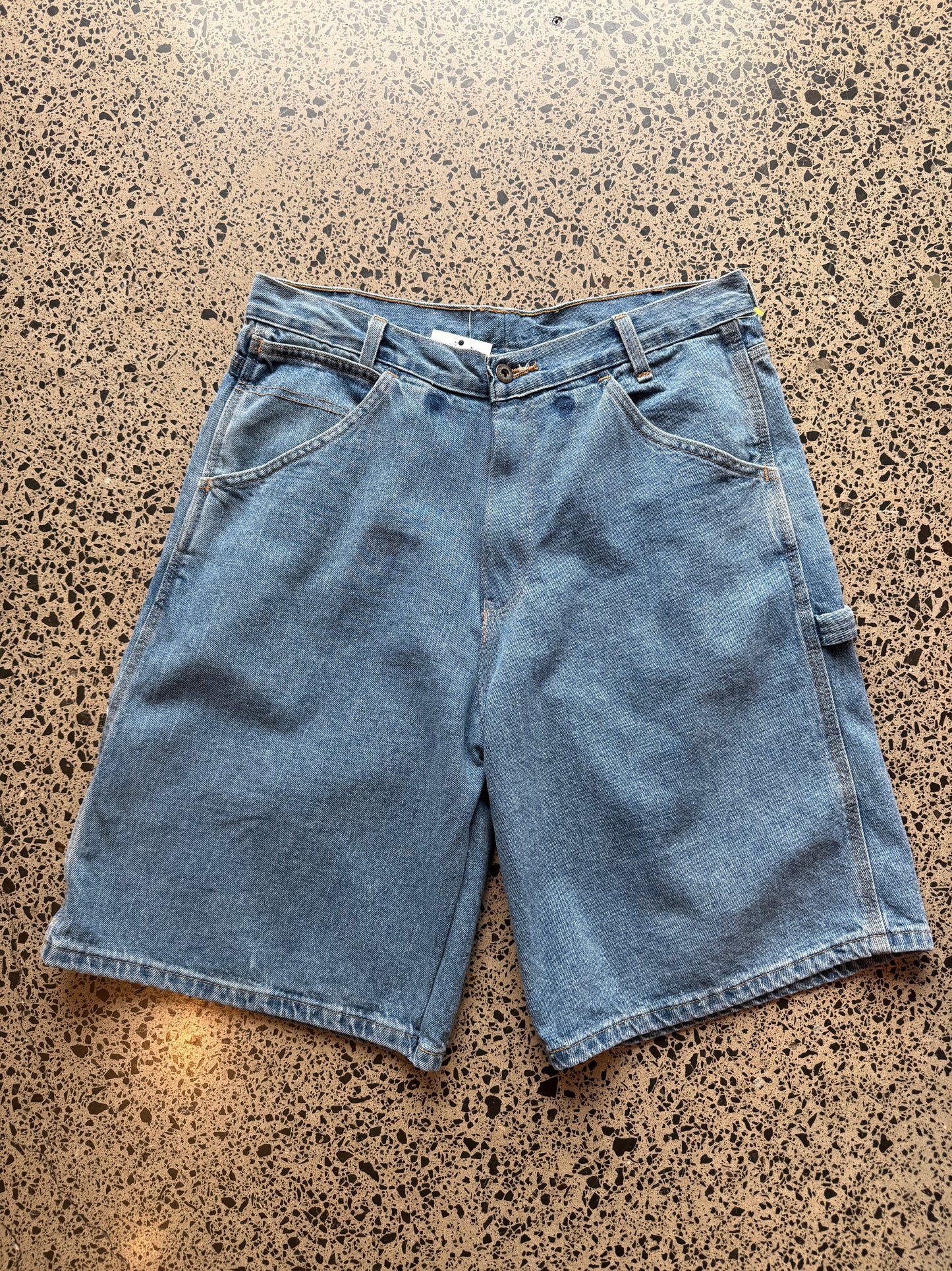 Light - mid blue wash denim jorts, size 32W, laid on polished concrete background. Knee cut with classic stitching and soft wash