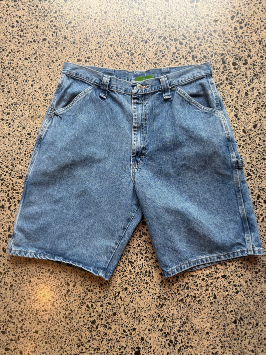 Wrangler Original mid blue wash denim jorts, size 32W, laid on polished concrete background. Knee cut with classic stitching and soft wash