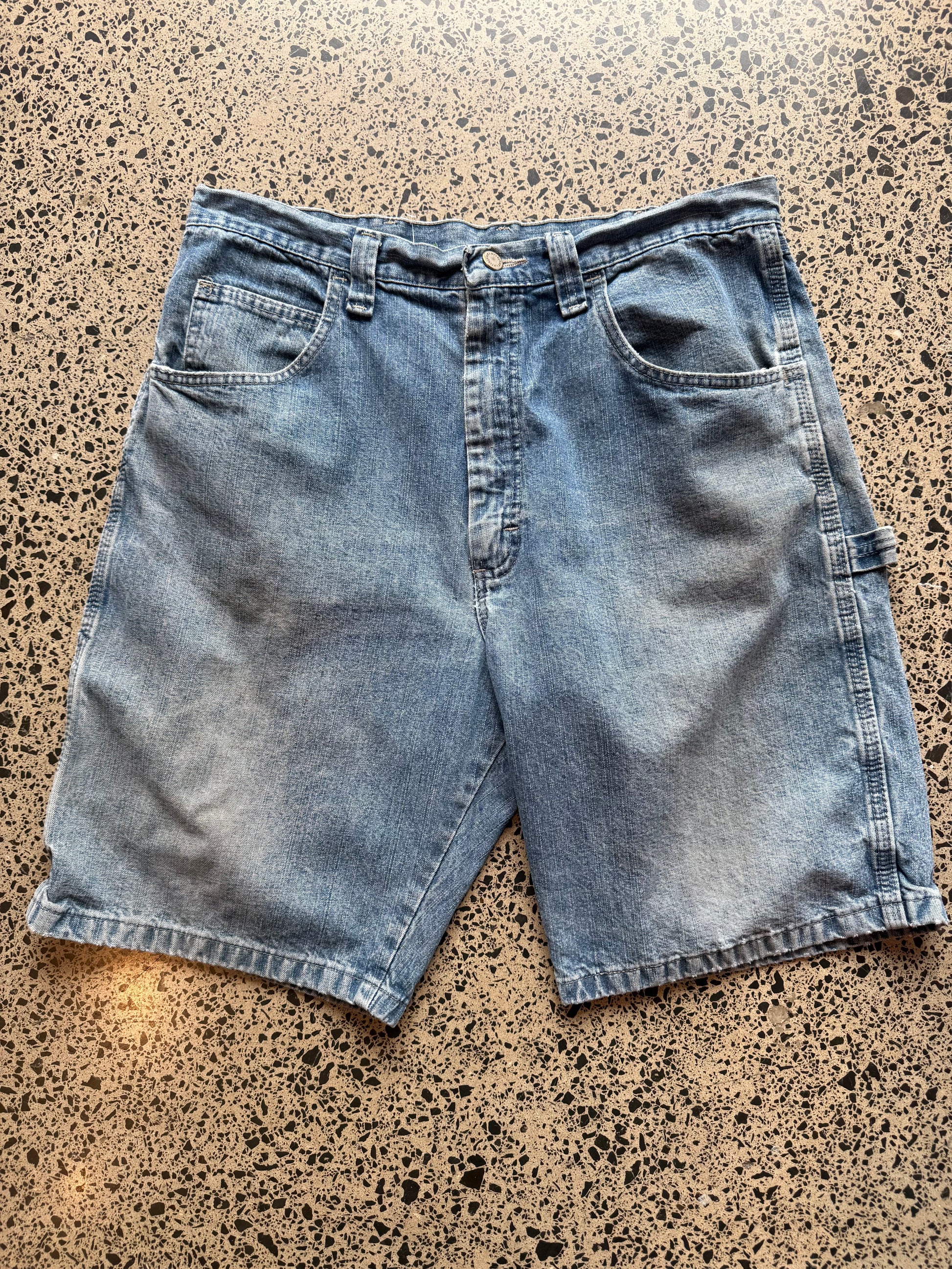 Wrangler Original light blue wash denim jorts, size 34W, laid on polished concrete background. Knee cut with classic stitching and soft wash