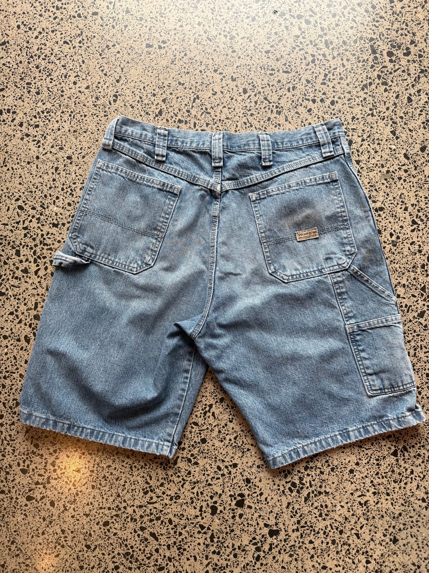 Wrangler Original light blue wash denim jorts, size 34W, laid on polished concrete background. Knee cut with classic stitching and soft wash