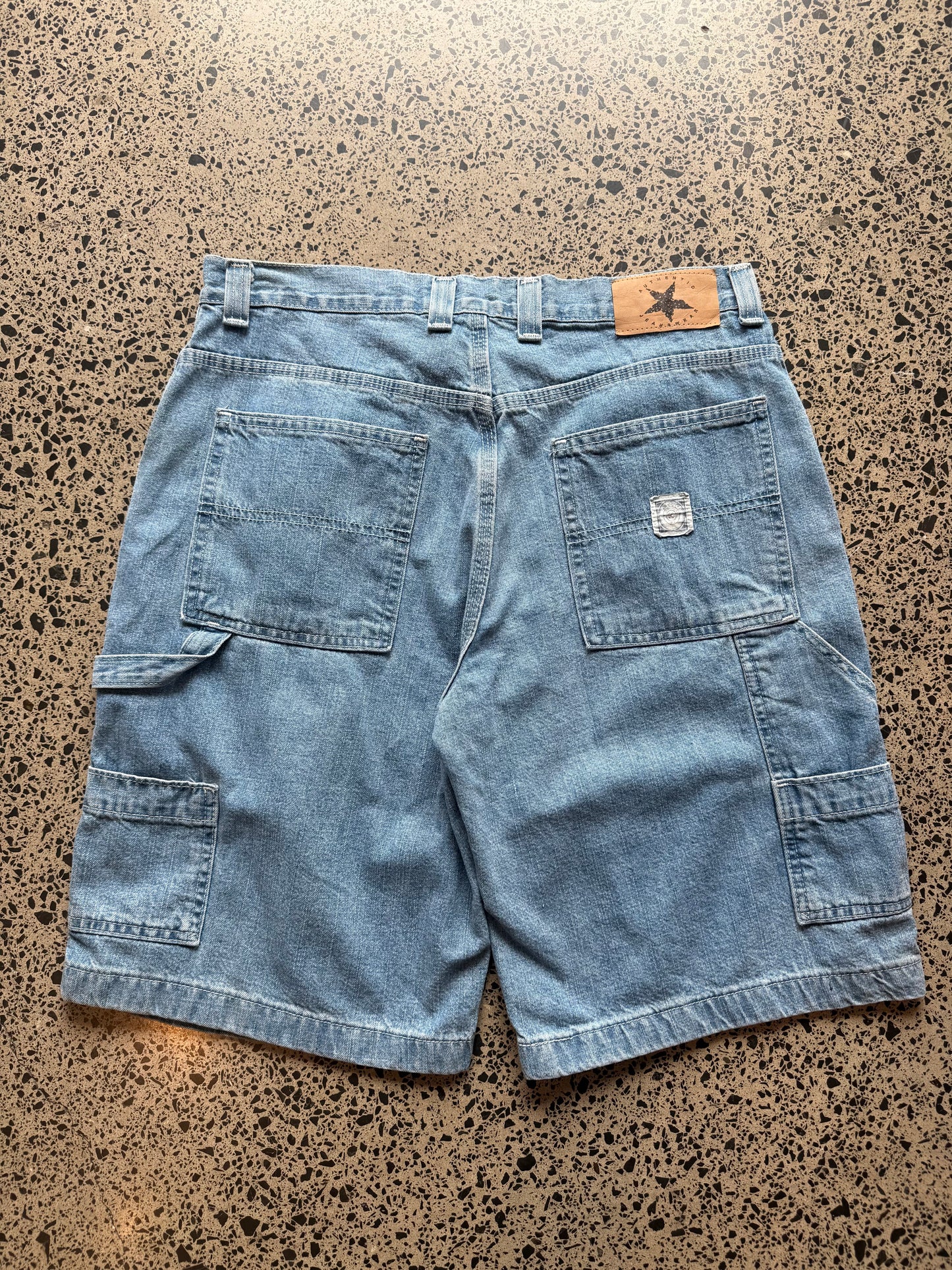 light blue wash denim jorts, size 34W, laid on polished concrete background. Knee cut with classic stitching and soft wash