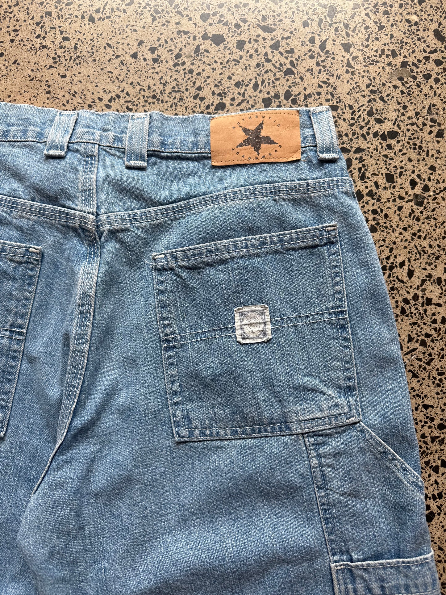 light blue wash denim jorts, size 34W, laid on polished concrete background. Knee cut with classic stitching and soft wash