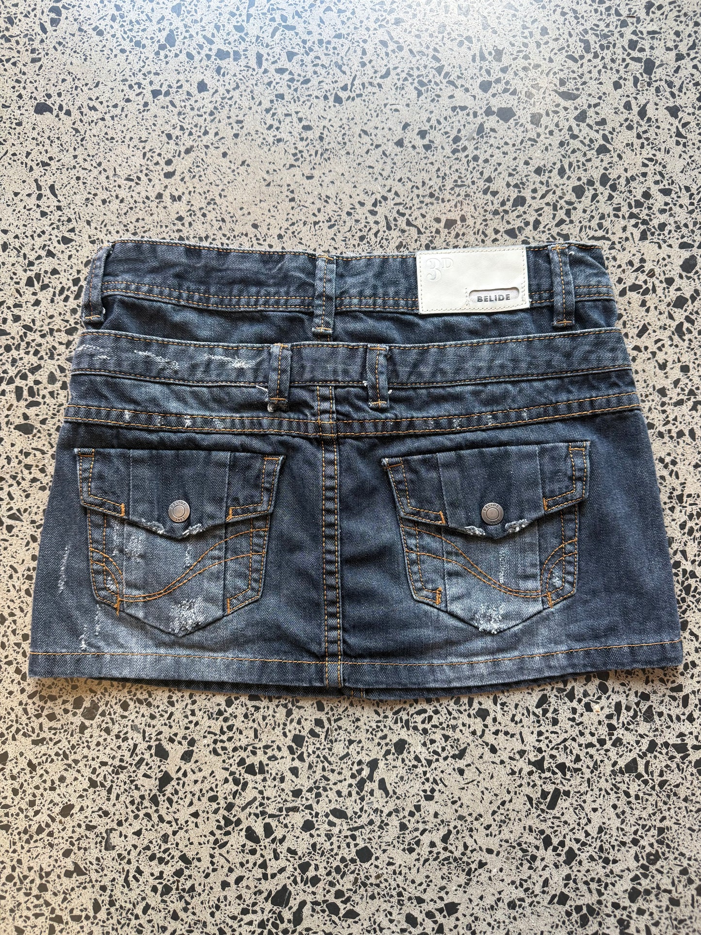 Y2K dark-wash denim mini skirt in XS laid on a polished concrete background — A-line cut with classic stitching and raw hem details.