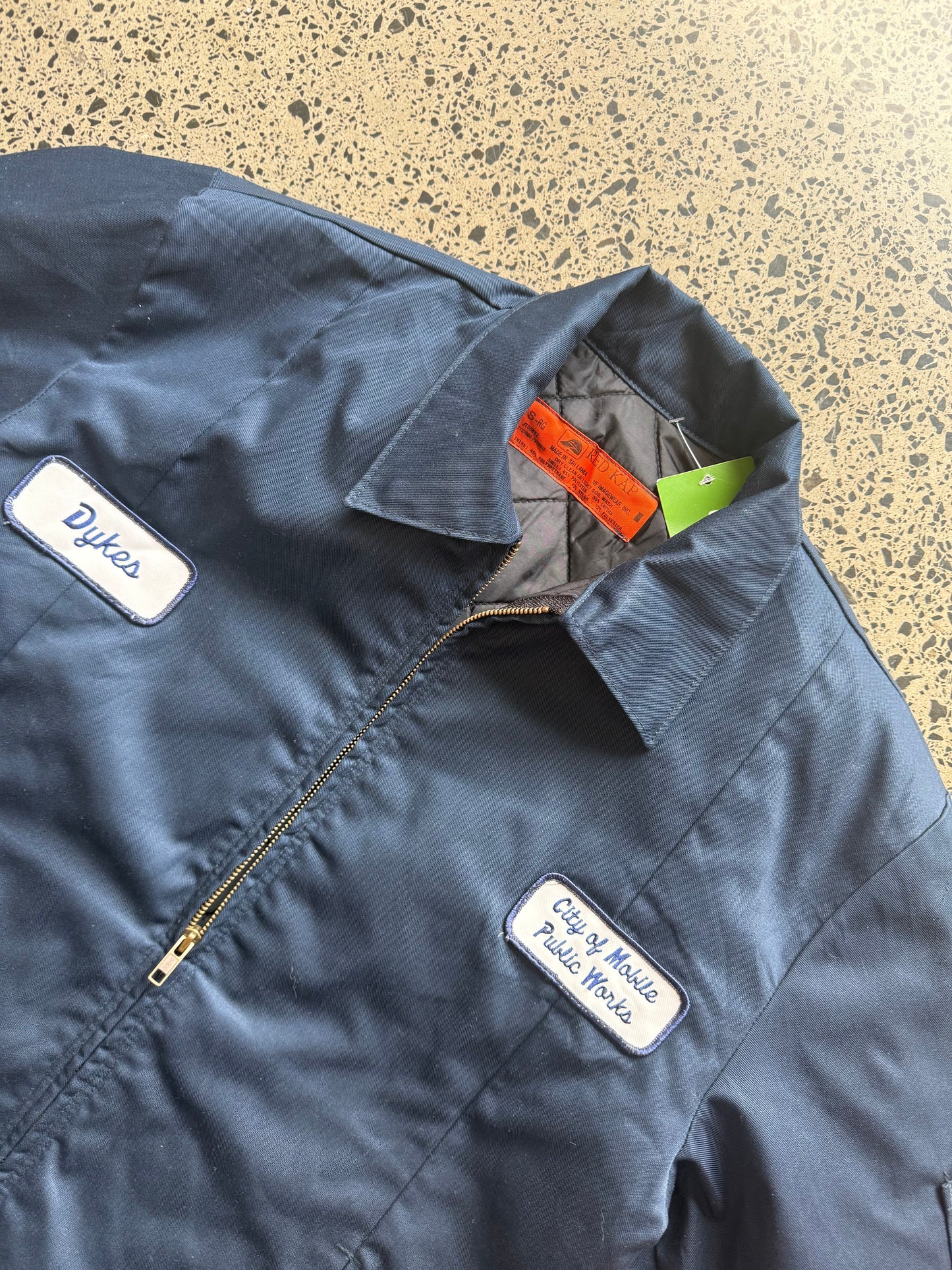 Red Kap workwear zipper jacket size Small Regular laid on polished concrete background — dark navy with zip front and pockets.