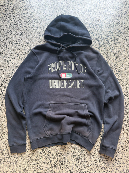 2010s Nike Undefeated hoodie size M laid on polished concrete background — black with bold graphics.